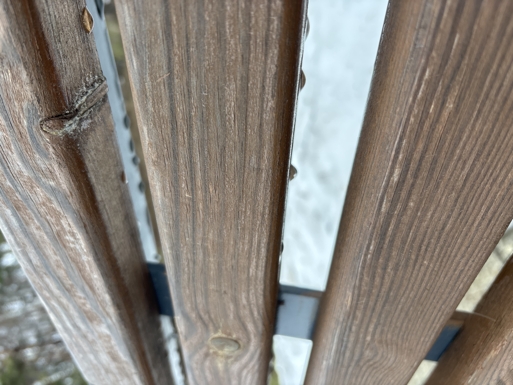 The image depicts a close-up, low-angle view of a wooden fence with gray metal supports. The wood has a brown, weathered appearance with visible wood grain and knots. Water droplets are present on the surface of the wood. The background shows blurry white and green areas, possibly snow or foliage. The location, Chaikovsky, Russia, is not discernible from the image.