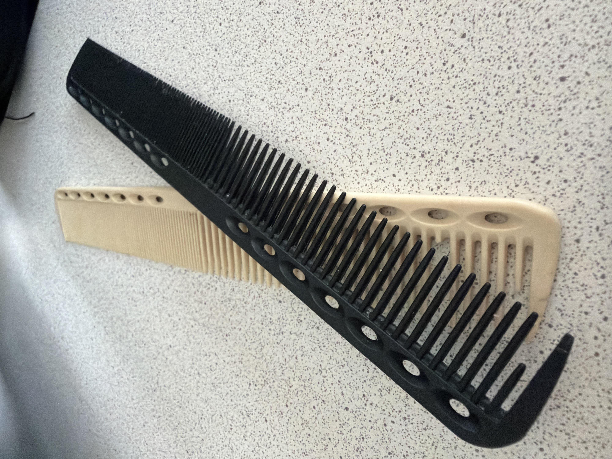 Two combs are resting on a speckled surface. One comb is black, with a series of circular cutouts along its spine and a curved end. The second comb is a pale beige color, and it lies underneath the black comb. There are no people visible in the frame. The environment is a surface with a fine, speckled pattern. The image does not contain sufficient information to determine the location in the city of Pervomaysky, Russia.