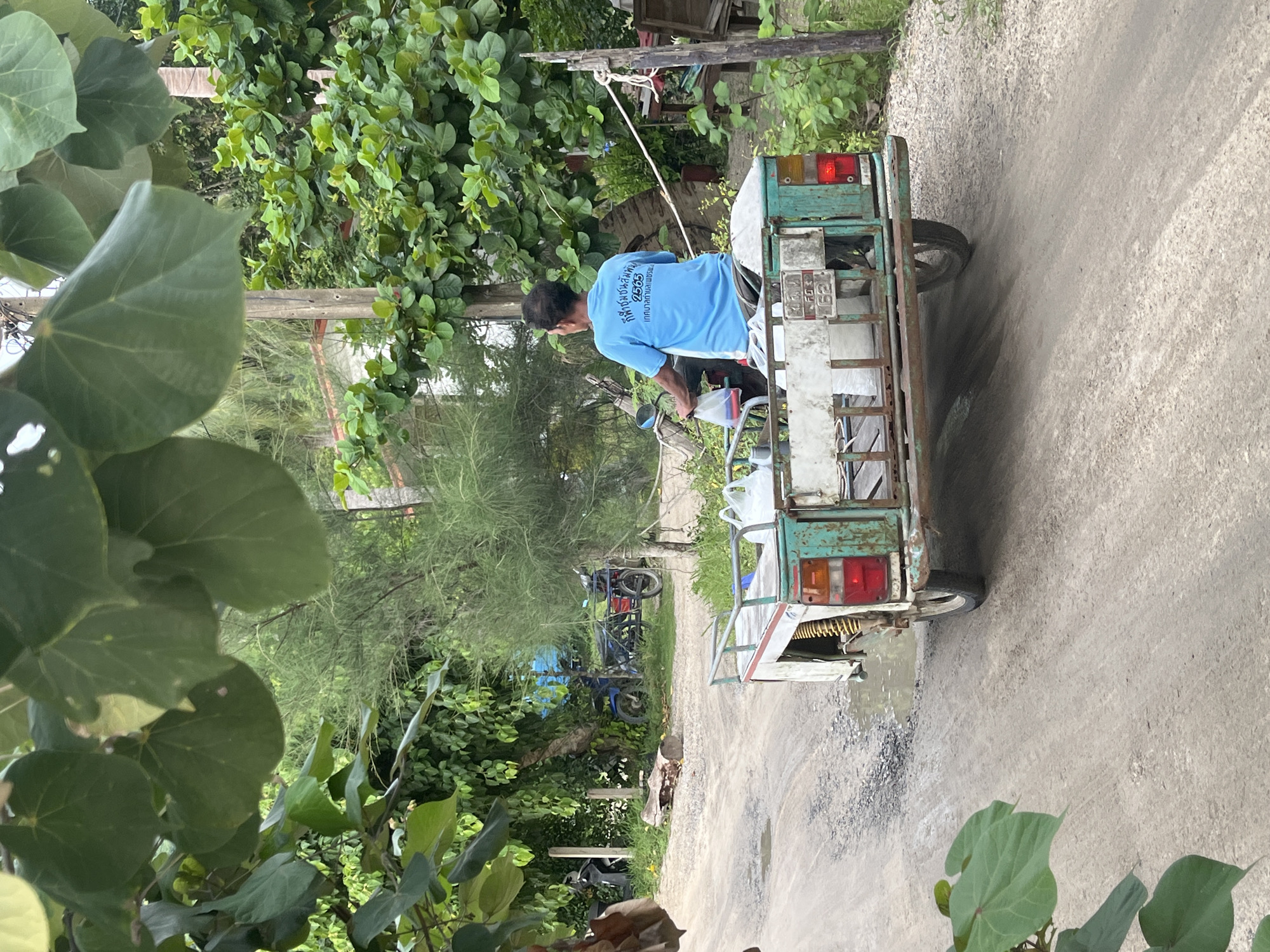 An elderly Asian man wearing a blue t-shirt is riding a three-wheeled vehicle on a dirt road in Ko Pha Ngan, Thailand. The vehicle is green and white, with a trailer attached to the back carrying several large white containers. The man is holding the handlebars and looking forward. The road is unpaved and has some puddles. In the background, there are trees and some buildings. The scene appears to be in a rural or semi-rural area.