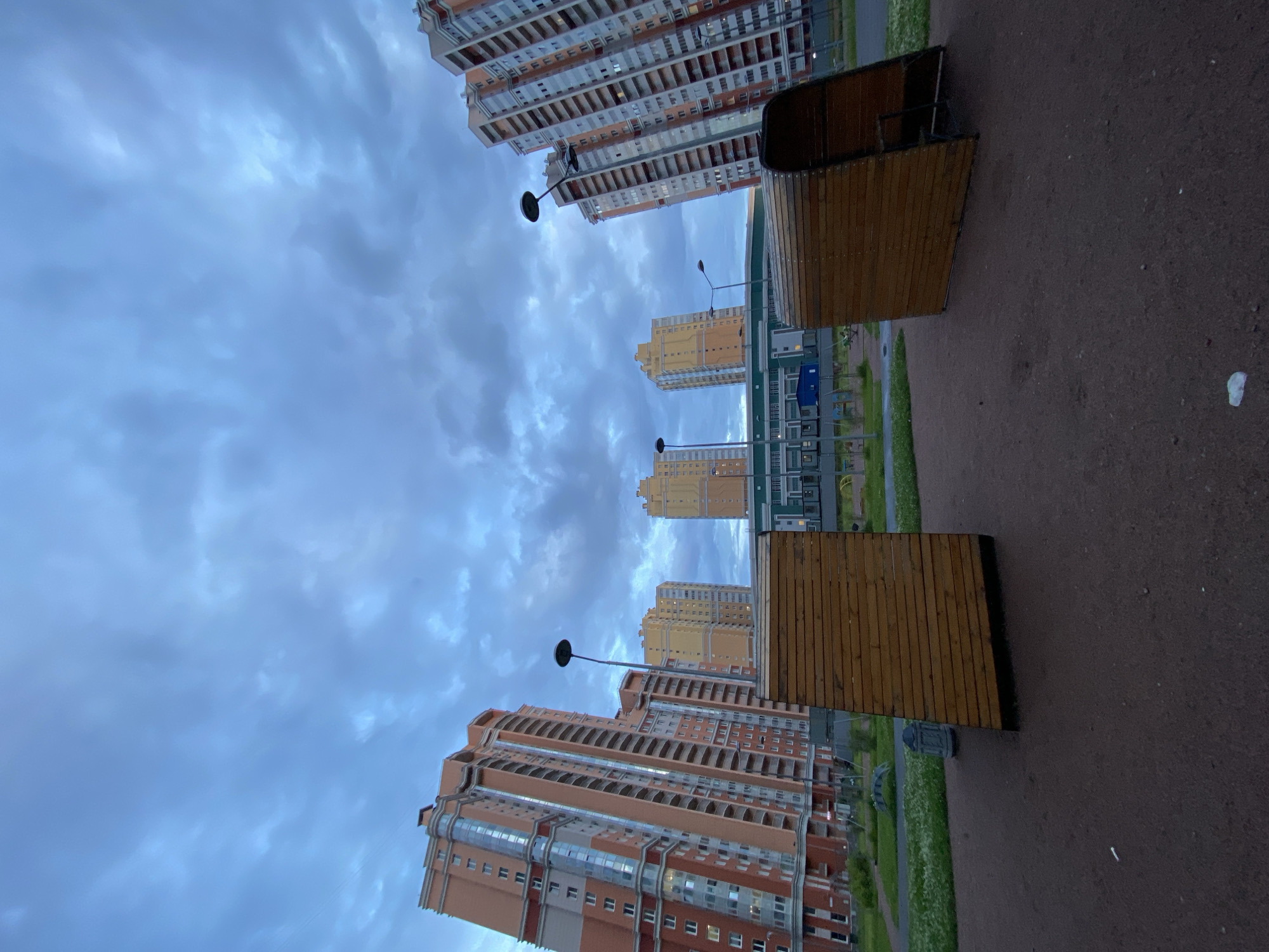 In an urban outdoor setting under a cloudy, overcast sky, two angled wooden structures, resembling benches or shelters, are positioned on dark brown gravel. Beyond the gravel path, a grassy area with scattered white flowers is visible, followed by a row of tall, modern residential buildings in shades of orange and yellow. Streetlights stand in the middle ground between the wooden structures and the apartment buildings. The architecture of the buildings features multiple floors with numerous windows and balconies.