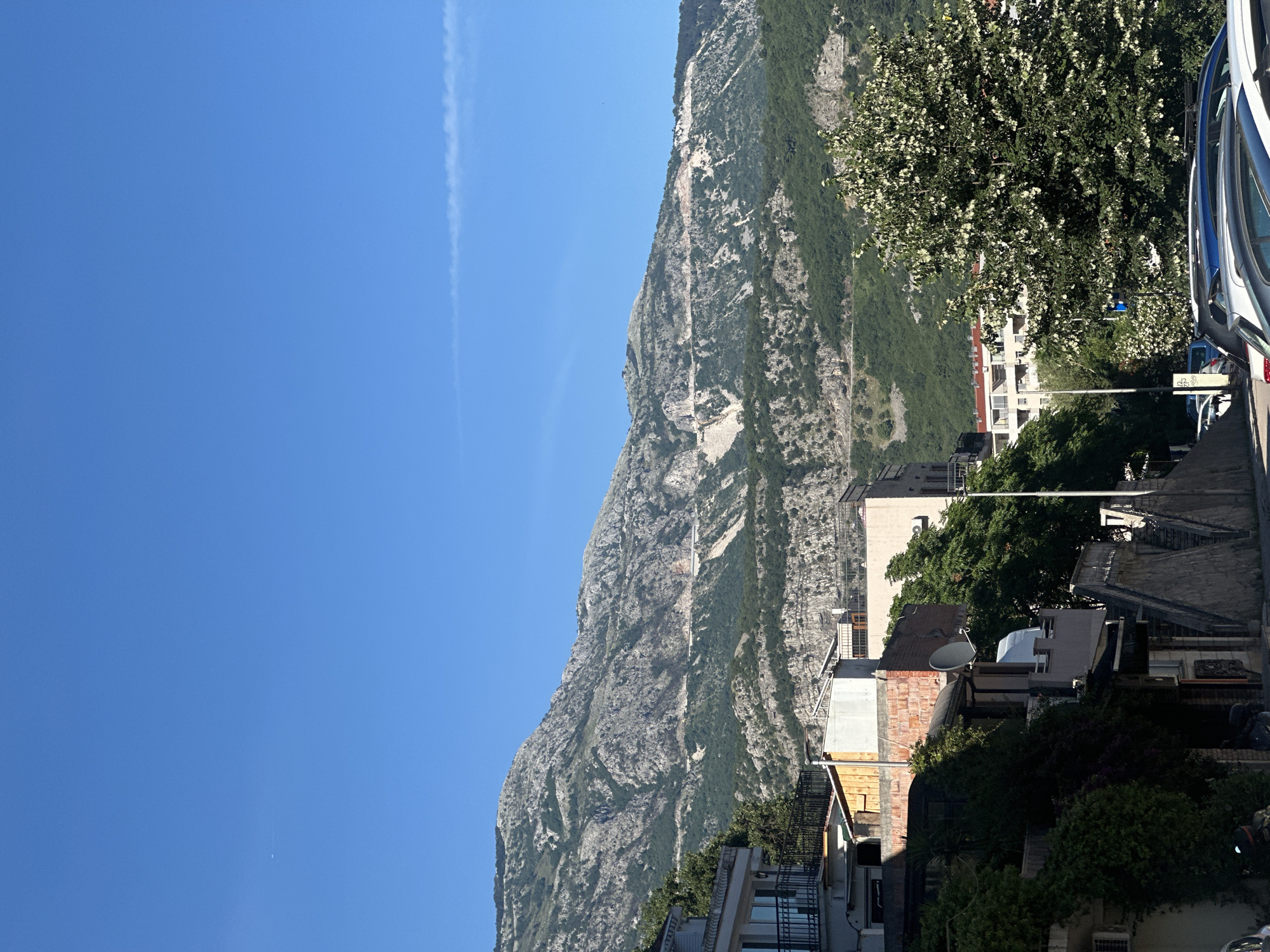 The image displays a bright, sunlit outdoor scene, likely during the daytime, situated in an urbanized area at the base of a mountain range, consistent with the city of Budva, Montenegro.
The upper portion of the frame is dominated by a clear, intensely blue sky. A faint, linear white contrail stretches horizontally across the upper right quadrant. A minuscule white object is discernible in the upper left section of the sky.
The background features a large, rugged mountain range, characterized by exposed grey rock faces and interspersed patches of dense green vegetation, primarily trees and shrubs. A distinctive winding road or track traverses the mountainside, evident by its lighter, excavated surface cutting through the rock and foliage.
In the mid-ground, an assortment of multi-story buildings is visible. On the far left, a light-colored building with dark-railed balconies is partially covered by vigorous green climbing plants. Towards the center, structures with varying finishes, including red brickwork, white plaster, and flat rooftops, are present; one building features a white satellite dish. Further to the right, behind a prominent tree, additional residential-style buildings with light facades are partially obscured.
The foreground depicts a street area. Multiple parked passenger vehicles, including sedans and hatchbacks of silver, blue, and dark colors, are aligned parallel to the street and facing the right, occupying the lower right quadrant. A portion of a motorbike is visible behind a parked car on the far left. A tall, slender street light pole ascends from the street level near the center. A large, leafy tree with light-colored clusters of blossoms or new growth dominates the mid-right foreground, partially obscuring the street and adjacent structures. No human individuals are visible within the depicted scene.