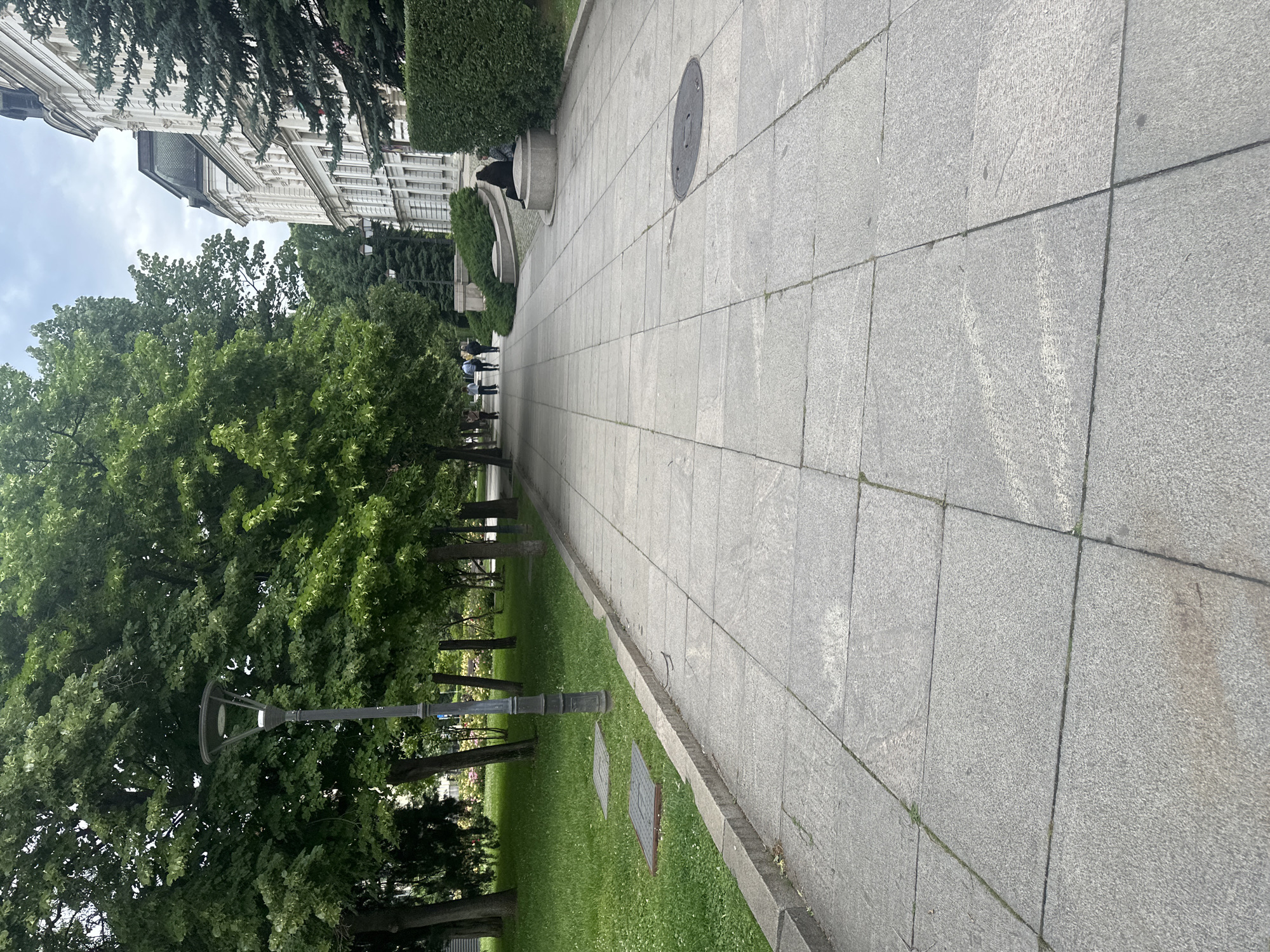 The image shows a pathway in what appears to be a park or garden setting. A stone-paved walkway stretches into the distance, with grass and trees on the left. A building can be seen on the left as well. The location is in Sofia, Bulgaria. People can be seen further along the walkway. A lamppost stands on the left side of the walkway. There are rectangular metal hatches are in the grass along the walkway.