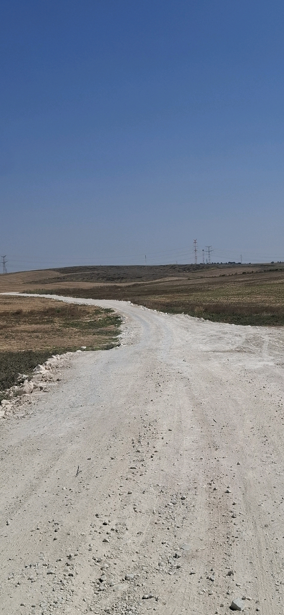 The image depicts an outdoor scene with a light-colored dirt road curving through dry, barren fields under a clear blue sky. The road appears to be unpaved, with visible tire tracks and scattered small rocks. The surrounding landscape consists of dry grasses and some patches of greenery, with rolling hills in the background. Several tall electricity pylons are visible on the horizon, suggesting the presence of power lines stretching across the landscape. The overall environment appears to be arid or semi-arid. The image does not contain any people or specific landmarks that would indicate its precise location within Kiryat Gat, Israel.