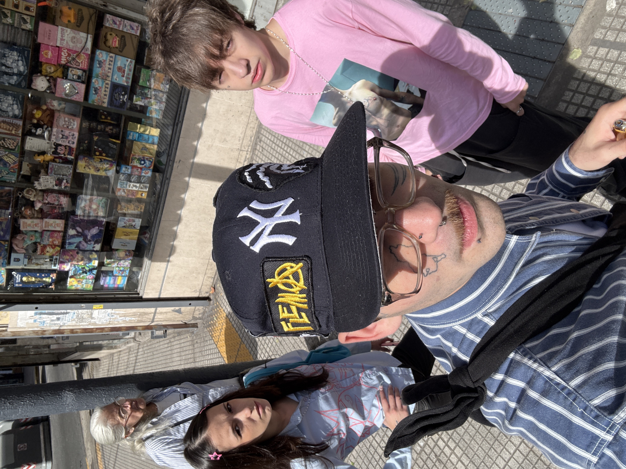 A medium shot captures a group of four individuals, seemingly posing for a photograph. The scene unfolds on a sunlit street, suggesting daytime. In the background, a storefront display of packaged goods is visible. The setting is the city of Buenos Aires, Argentina.
At the forefront of the frame, a person wearing a baseball cap and spectacles, sporting a mustache, and dressed in a blue and white striped shirt faces the camera. Adjacent to this individual, another person is wearing a pink long-sleeved shirt with a graphic design.
Behind these two individuals, there are two additional people. A woman with long dark hair is facing the camera, and behind her, a person with short white hair is facing the camera.
The composition is taken from a slightly elevated perspective, looking down at the subjects, adding depth to the overall composition.