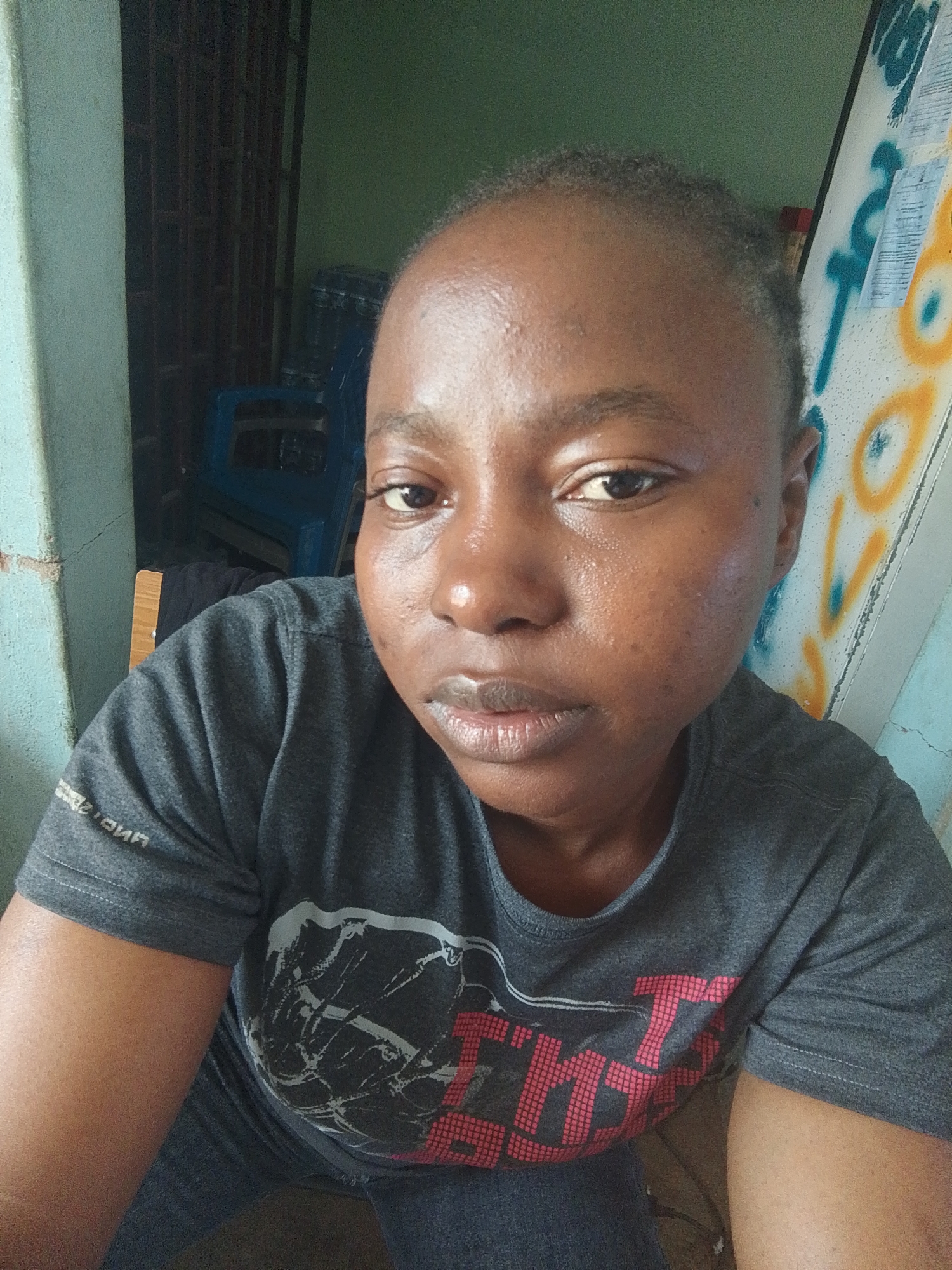A waist-up, close-up selfie captures a Black individual, likely female, with short, dark, natural hair, looking directly at the camera with a neutral expression. Her dark skin tone shows some natural texture and a slight sheen, possibly from natural oils or lighting. She is wearing a dark grey short-sleeved t-shirt with a graphic design featuring white abstract elements and bold red text on the chest, where the word "FIT" is clearly discernible.
The setting appears to be an indoor space in Jos, Nigeria, characterized by varied wall finishes and ambient lighting. To the left of the subject, a textured, light blue-green wall or pillar is visible, alongside a darker interior area that includes part of a wooden chair frame and a blue plastic crate or container. To the right, a white wall is adorned with abstract graffiti-like patterns in blue and yellow, featuring circular and linear shapes. The lighting suggests it is daytime. The image conveys a casual and candid moment.