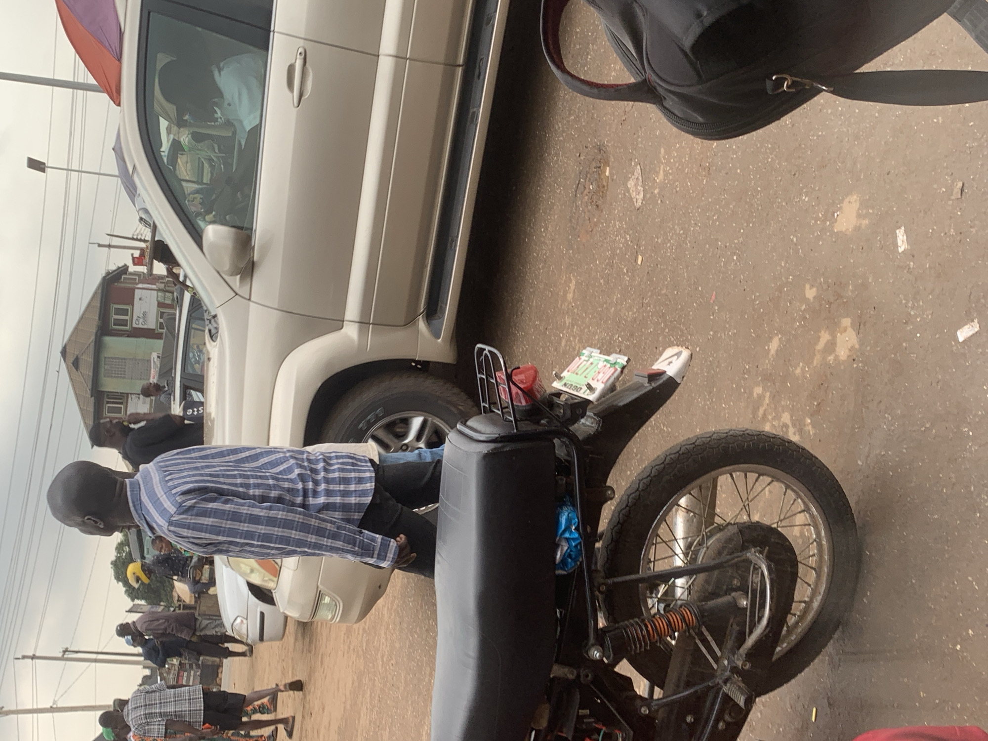 In Shagamu, Nigeria, a light-colored SUV is parked on a dirt road. The driver, a man with dark skin and short hair, is visible inside the vehicle, wearing a white shirt. In the foreground, a black motorcycle is parked, facing right. A person wearing a blue and white striped shirt and dark pants is standing near the motorcycle, looking away from the camera. Other individuals are present in the background, some standing, and one riding a motorcycle with a yellow helmet. Buildings with visible windows and roofs are in the distance, suggesting an urban or semi-urban environment. The ground is composed of reddish-brown dirt and gravel. The sky is overcast.