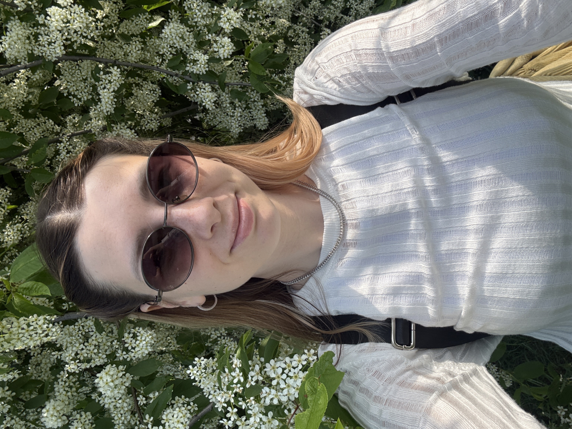 A woman wearing sunglasses, a white long-sleeved shirt, and a silver necklace is lying among white flowers and green leaves. She has brown hair with blonde highlights. A black strap, possibly from a bag, is visible across her torso. The location appears to be outdoors, possibly a park or garden setting. The precise location within Moscow, Russia, cannot be determined from the visual information provided.