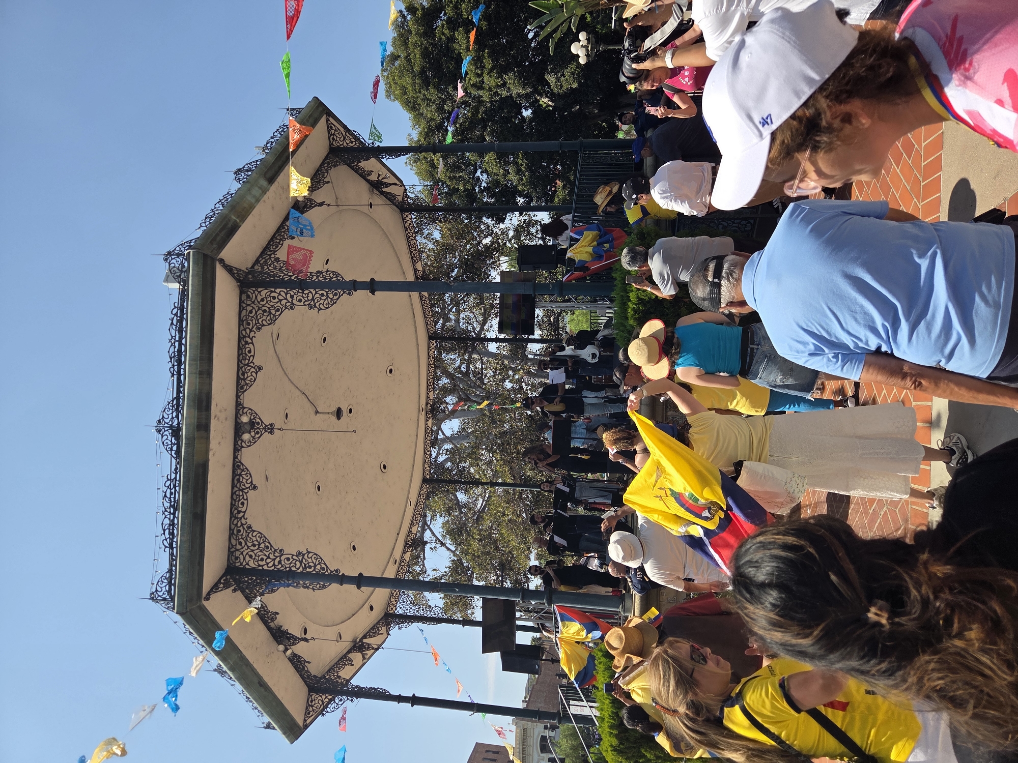 This image depicts an outdoor gathering at a public venue, possibly a park or plaza, featuring a decorative gazebo or bandstand adorned with colorful paper flags. A band is performing on a stage set up within or in front of the gazebo. A crowd of people is gathered, many holding up flags, including one prominent flag with yellow, blue, and red stripes. The overall atmosphere suggests a cultural event or celebration.
While the image itself does not contain explicit geographical markers, the presence of such a gazebo and the style of architecture, combined with the cultural themes (e.g., flags, potentially music), could suggest a location like Olvera Street in Los Angeles. Olvera Street is a historic street and marketplace in downtown Los Angeles, known for its vibrant Mexican culture, historic buildings, and frequently hosting cultural events and festivals.