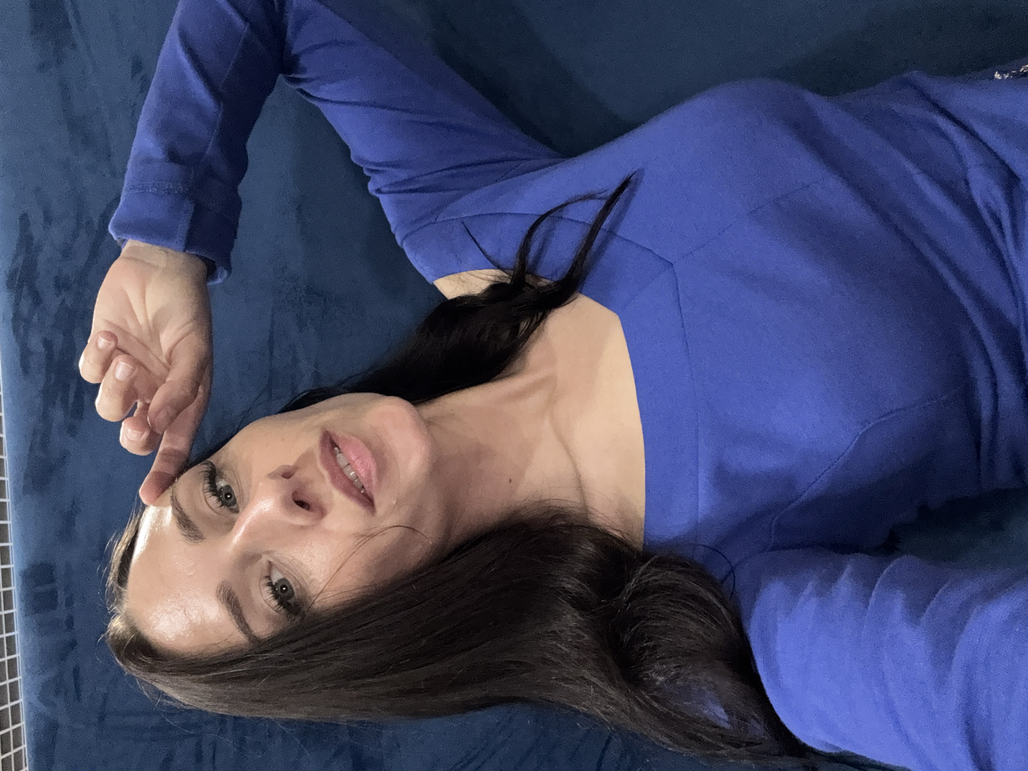 A woman with long, dark hair and light-colored eyes is captured in a close-up selfie taken indoors. She is wearing a royal blue long-sleeved top with a V-neckline and cut-out shoulder details. Her right hand is raised to her forehead, touching her brow with her index finger. The background appears to be a textured, dark blue surface, possibly a couch or upholstery. The image is a medium shot, focusing on the woman's face and upper body. There is no discernible connection to Saint Petersburg, Russia, or any specific city location.