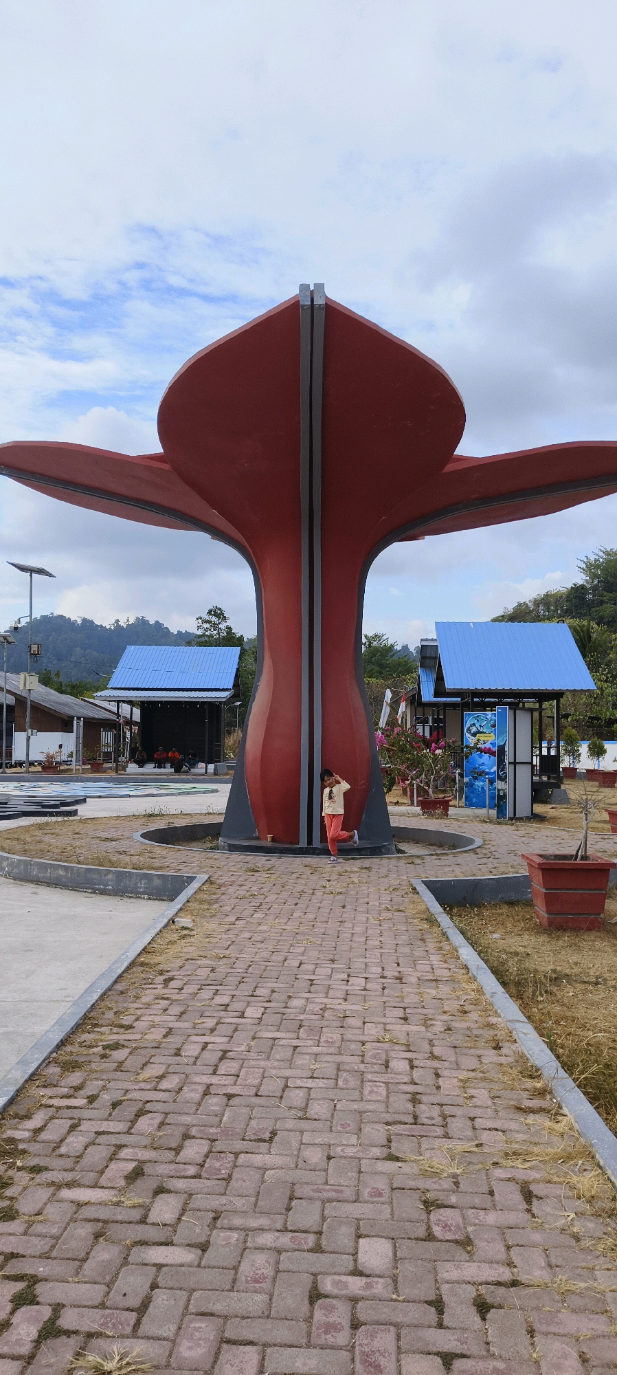 The image depicts a public park area in the city of Waisai, Indonesia, featuring a large, red, petal-shaped sculpture as its central element. A child, wearing a light-colored long-sleeved shirt and orange pants, is standing to the right of the sculpture, posing with their hand raised near their head. The ground in the foreground is paved with reddish-brown bricks laid in a herringbone pattern, bordered by concrete pathways and dry grass.
Behind the sculpture, two small structures with blue roofs are visible. The structure on the left appears to be a covered seating area or market stall, with several people seated inside. The structure on the right is a kiosk or information booth, featuring several posters with marine-themed imagery. In the background, lush green hills covered with trees form the horizon, under a cloudy sky. Solar panels are visible on a pole to the left of the sculpture, suggesting modern infrastructure within the park. The overall scene conveys a sense of an outdoor recreational space with distinct architectural and natural elements.