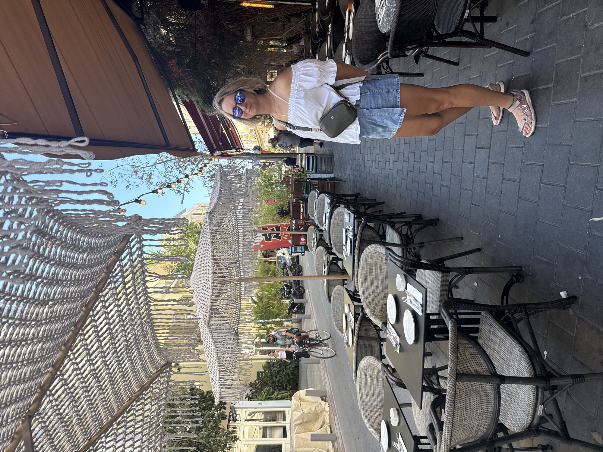 A woman stands on a sidewalk lined with outdoor cafe tables in Tel Aviv, Israel. The woman is wearing a white off-the-shoulder top, a denim skirt, and pink sandals. She has sunglasses on and is carrying a dark green crossbody bag. The cafe has tables and chairs set up on the pavement, with umbrellas providing shade. In the background, people are walking and cycling on the street, and there are motorcycles parked. The architecture suggests an urban environment with buildings on either side of the street. The overall scene depicts a pleasant day at an outdoor dining establishment in a city.