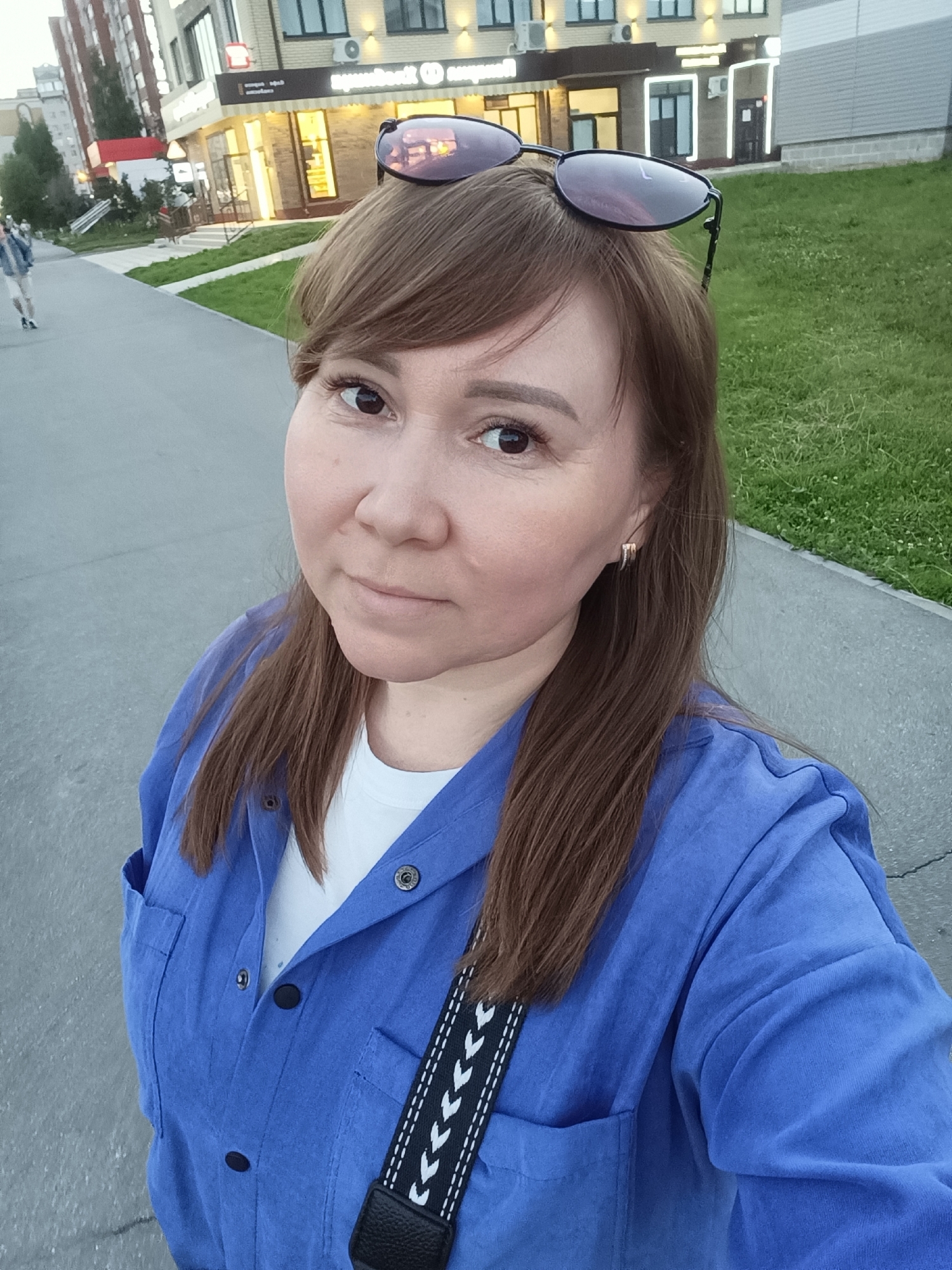 A woman is taking a selfie outdoors. She has medium-length brown hair and is wearing sunglasses on top of her head. Her eyes are brown, and she is wearing a blue shirt over a white t-shirt. A black strap with a white chevron pattern is visible across her chest. She is standing on a paved path next to a grassy area. In the background, there is a modern building with large windows and illuminated signage. Further in the distance, another building and some trees are visible. The lighting suggests it is either early morning or late afternoon. The weather appears to be clear. The overall scene depicts a casual outdoor moment in a developed urban or suburban area.