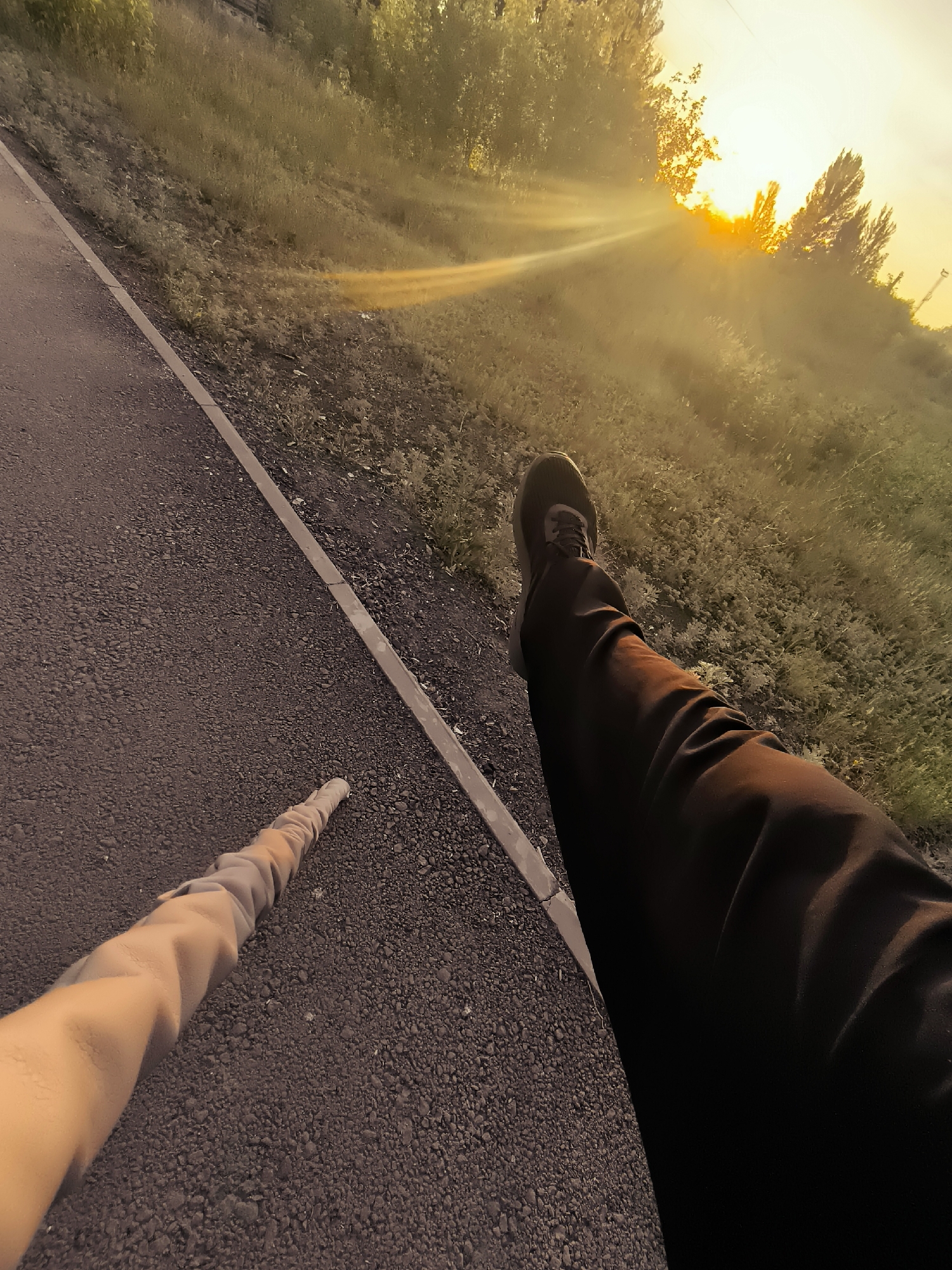 The image shows a first-person perspective of someone's legs and arm from the knees down, positioned on a paved path next to a grassy slope. The sun is low in the sky, casting a warm, golden light and creating visible rays through a slight haze or mist. Trees are visible on the slope, silhouetted against the bright sun. The pavement appears to be asphalt with a concrete curb. The person is wearing dark pants and dark athletic shoes on their right leg, which is extended. The left arm, with a light-colored sleeve, is also visible, extended forward and slightly downward. The overall impression is of an outdoor scene during sunrise or sunset, likely in a park or along a pathway within Qarağandı city administration, Kazakhstan, given the context provided.