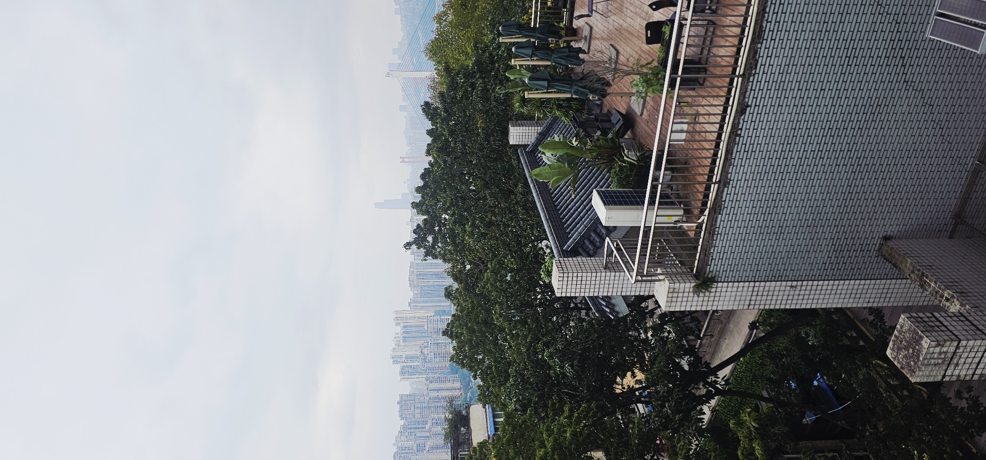 The image shows a view of a city from an elevated position, likely a balcony or rooftop. The cityscape is visible in the distance, obscured by haze and foliage. The right side of the image features a building with tiled surfaces, a balcony with a metal railing, and various architectural elements like eaves and rooftops. The immediate foreground contains greenery, including trees and foliage. The overall impression is an urban scene with a mix of natural and built environments. The location is identified as Shapingba District, China.
