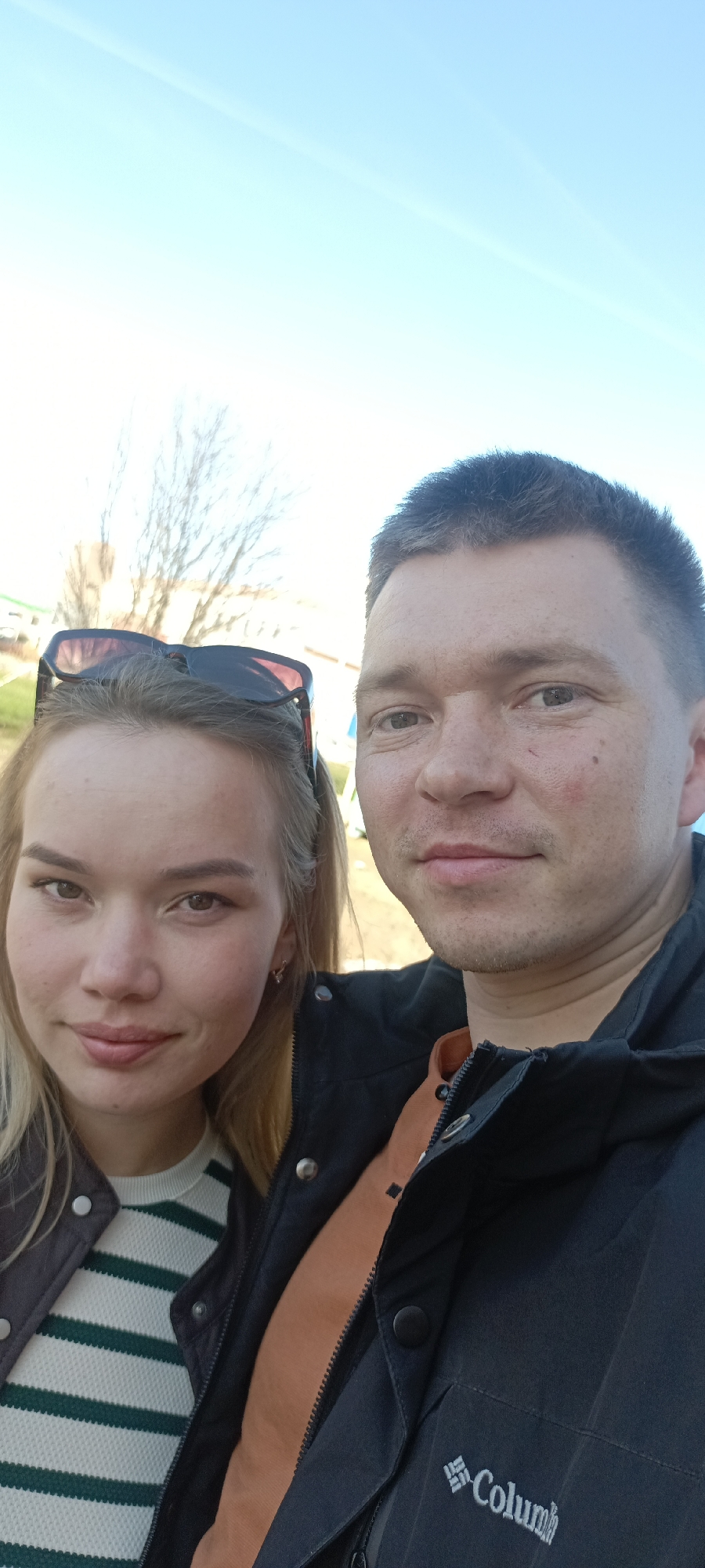 A close-up outdoor selfie captured in Markovskii, Russia, features two individuals under a bright, clear sky. On the left, a woman with fair skin and light-colored hair looks towards the camera with a gentle, slight smile. She has dark sunglasses resting on top of her head and wears a white sweater with horizontal green stripes, layered under a dark jacket. To her right, a man with short dark hair and a neutral to slightly smiling expression is visible. He wears a black outdoor jacket, beneath which an orange layer (possibly a shirt or jacket lining) is visible. The man's jacket prominently features a white logo with the partial text "Columb," indicative of the brand Columbia. The background, softly out of focus, shows bare tree branches, suggesting a non-summer season like late autumn, winter, or early spring, against a clear, light blue sky with subtle wisps of high-altitude clouds. The lighting is bright and natural, indicating daytime.