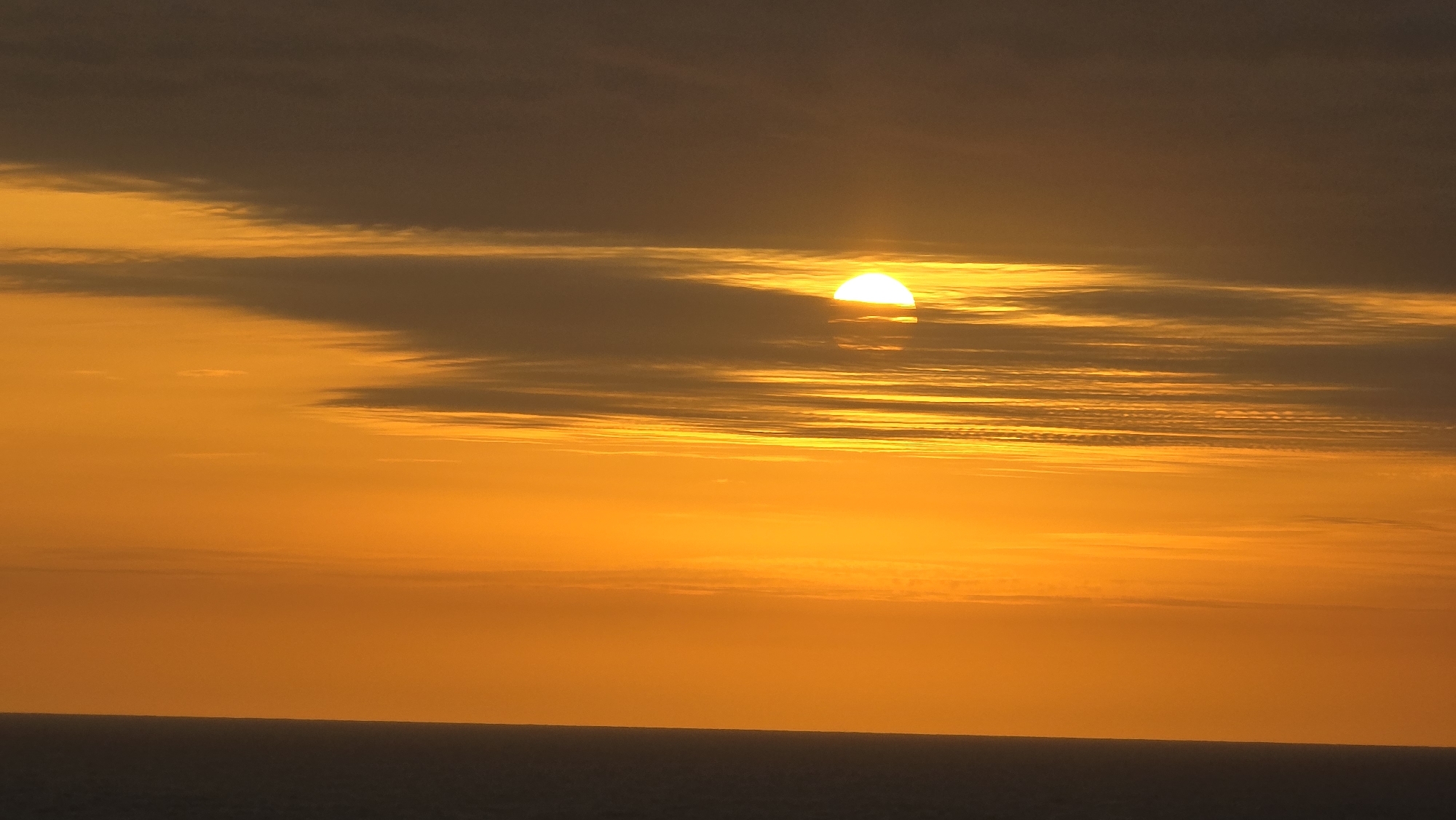 The image depicts a sunset over the sea, potentially taken in Randaberg, Norway. The sun is partially obscured by a thin layer of clouds, emitting a bright, golden light. The sky is layered with orange and gray clouds, becoming a deeper orange towards the horizon where it meets the dark sea. A flat, dark line indicates the horizon. No people are visible.