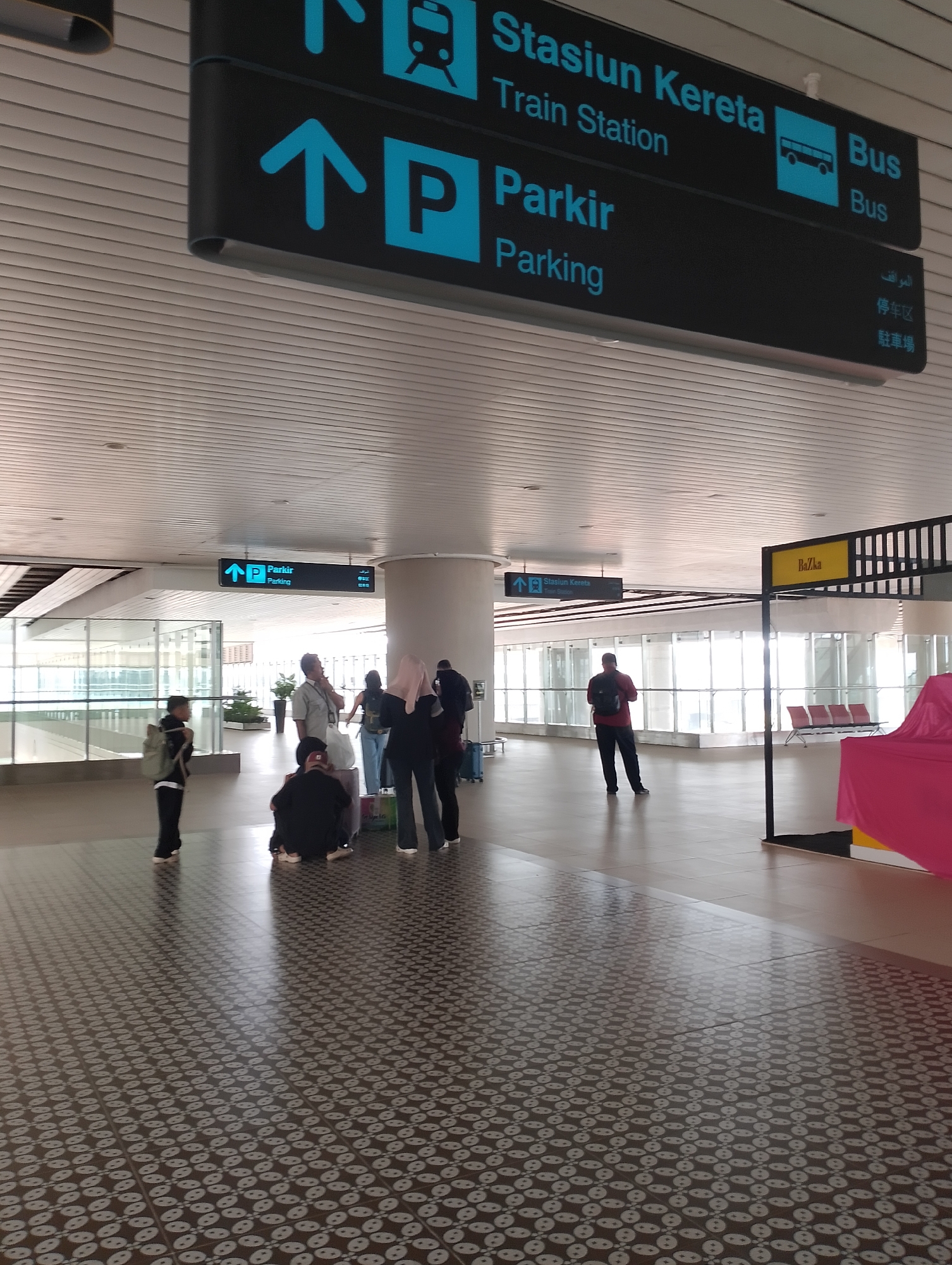 The image shows an indoor scene at a transportation hub, likely an airport or train station, in Palihan, Indonesia. Overhead, there are directional signs in blue and black. One sign indicates "Stasiun Kereta" and "Train Station" with a train icon and "Bus" with a bus icon. Another sign, further down, points to "Parkir" and "Parking" with a parking icon and directional arrows. There are also signs in what appear to be Arabic, Chinese, and Malay.
In the foreground and midground, several individuals are present. Some are standing and walking, while others are sitting on the floor or on a low bench-like structure. One person in the foreground is wearing a backpack. Another person, dressed in red, is standing with their back to the camera. There are groups of people interacting or waiting.
The environment features a modern ceiling with light panels. The floor is tiled with a patterned design. In the background, there are large windows with a view of what appears to be an outdoor area with seating. A yellow sign with the text "BaZka" is visible on the right side of the image, along with a structure covered in pink fabric. The lighting suggests it is daytime.