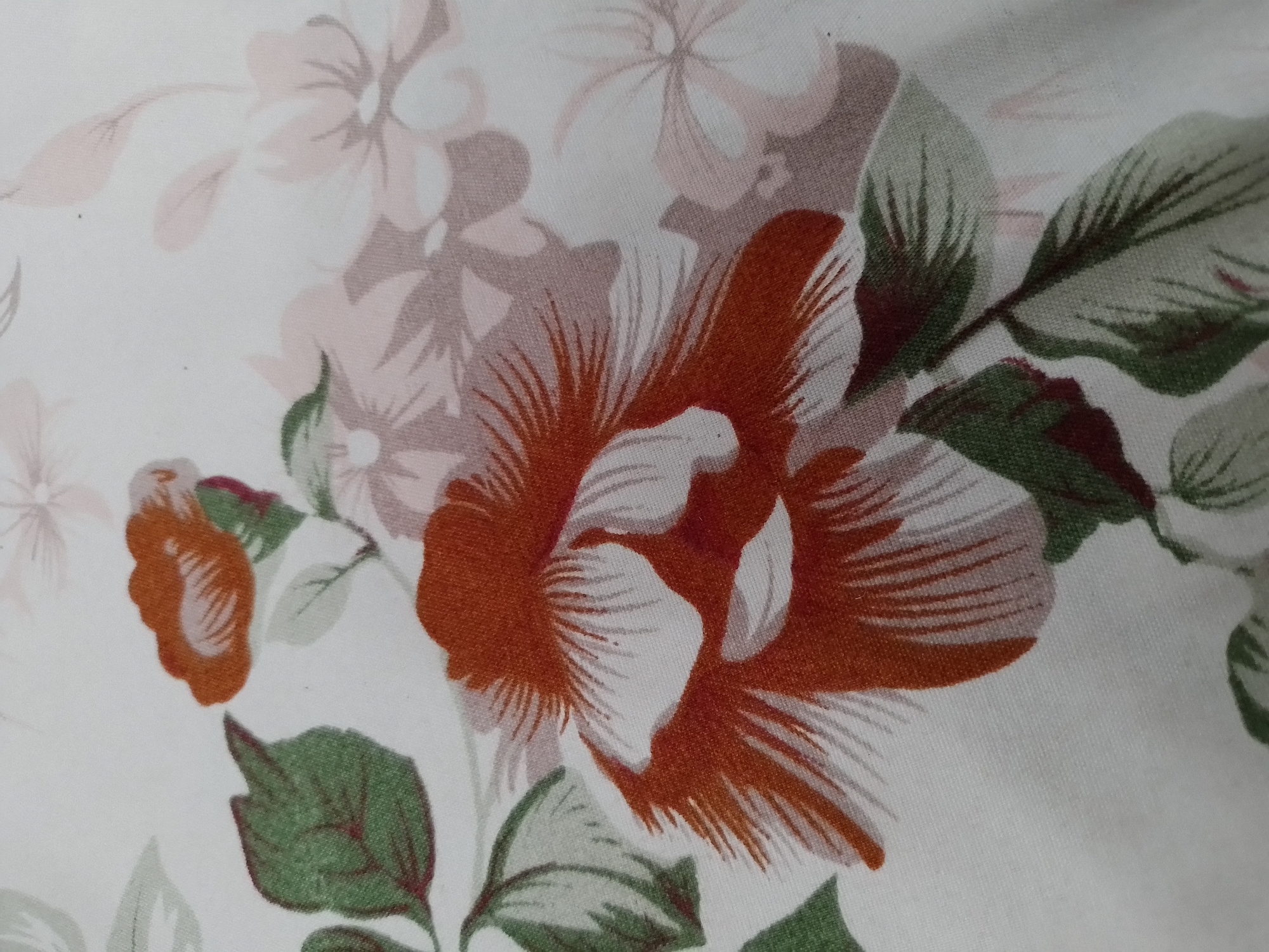 This is a close-up photograph of fabric with a floral pattern. The dominant feature is a large, stylized flower in shades of burnt orange and white, with a center of white petals radiating outwards. To the left of this flower are green leaves. The background is white with a subtle pattern of lighter, faded flowers and leaves in pink and grey tones. The fabric appears to be slightly textured. The image does not depict any specific location in Ngancar, Indonesia, nor does it contain any people or actions.