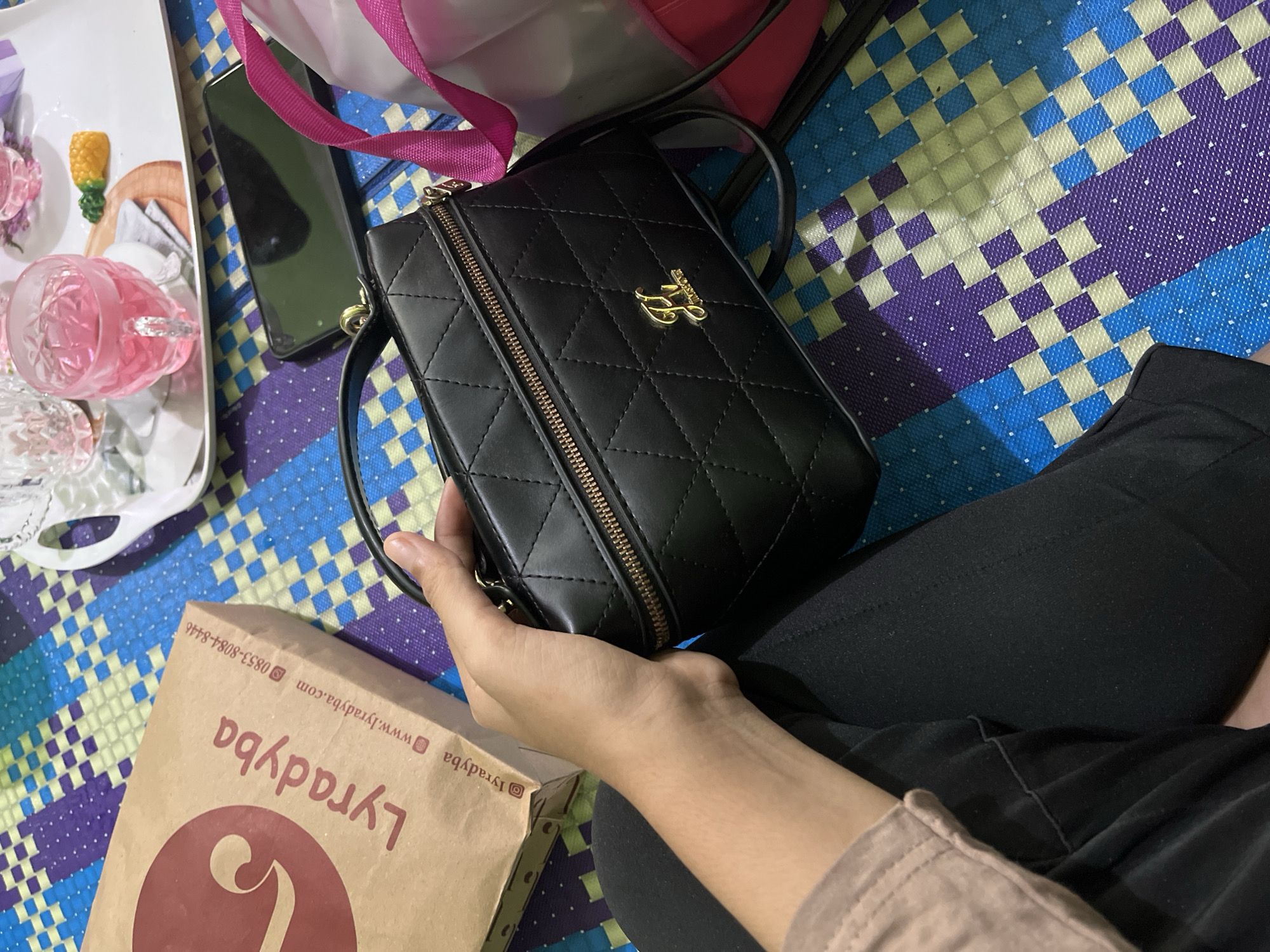 In Babahjurong, Indonesia, a person is holding a black quilted handbag with a gold logo. The handbag is positioned in the foreground, angled slightly to the right. To the left of the handbag, a brown paper shopping bag with the "Lyradbya" logo is visible. The background features a colorful, woven mat in shades of blue, purple, and yellow. A tray with two glasses of pink liquid, a small decorative pineapple, and a white napkin is placed on the mat behind the handbag. A smartphone lies on the mat to the right of the tray. A pink strap of another bag is partially visible to the right of the black handbag. The person's hand and arm, wearing a tan sleeve, are holding the handbag.