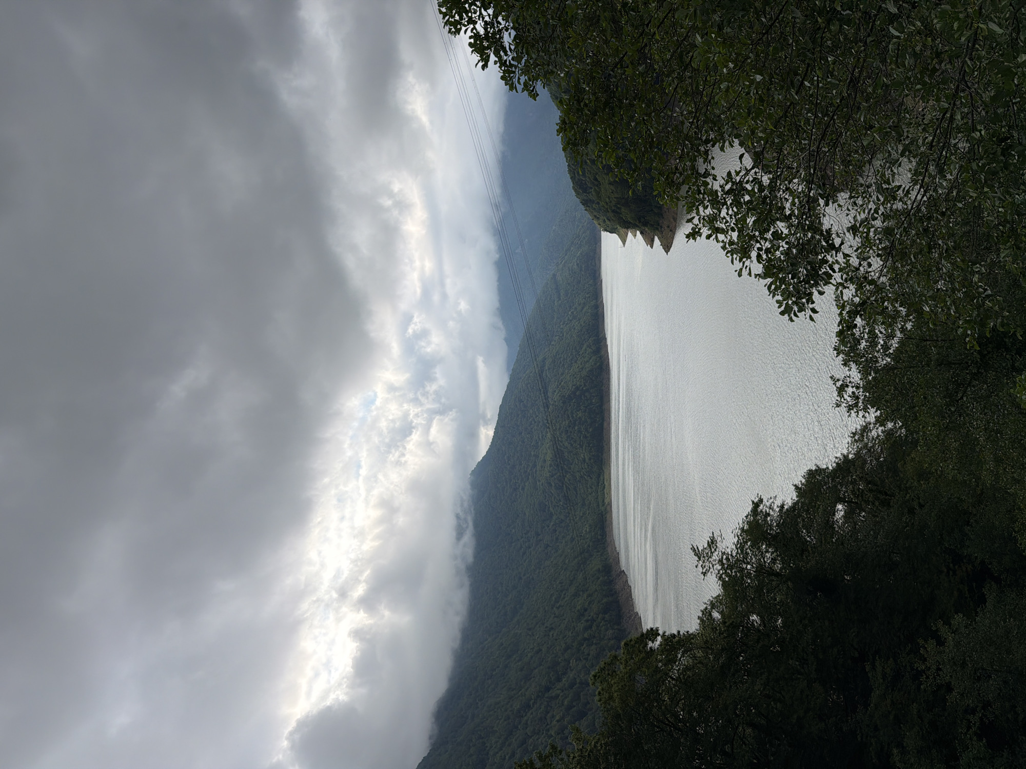 This image depicts a scenic view of a large body of water, likely a lake or reservoir, nestled beside a verdant, forested mountain range. The perspective is from a slightly elevated position, looking down towards the water and across to the mountains. The sky is overcast with dramatic, layered clouds, through which sunlight occasionally breaks, creating luminous patches and highlighting the textures of the clouds. Some of these clouds appear to be low-lying, hugging the contours of the mountains, adding a sense of depth and mystery to the landscape.
On the left side of the frame, the mountains are densely covered in lush green trees, their slopes rising steeply. The terrain appears rugged and untamed. Along the distant mountains, what appear to be power lines or cables are strung across, hinting at human infrastructure within this natural setting.
The body of water occupies the right half of the image, its surface rippled by a gentle breeze, reflecting the muted light from the sky. The water is a pale, silvery grey, contrasting with the darker greens of the mountains. In the foreground, on the right and top edges of the image, the dark silhouettes of leafy trees frame the view, their branches and foliage creating a natural border and adding a sense of intimacy to the expansive landscape.
The overall mood is one of serene, dramatic natural beauty. The overcast weather suggests it could be late afternoon or a moody, misty day. The location cues, such as the specific mountainous terrain and the presence of power lines, combined with the provided location context of Chveletana, Georgia, suggest a remote, natural area. There is no visible text in the image.
