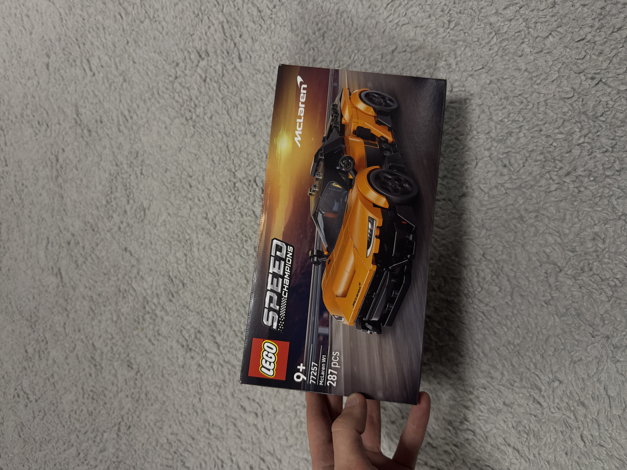 The image shows a Lego Speed Champions McLaren Solus GT and McLaren F1 LM set box. The box is held in a person's hand against a grey, textured background, likely a blanket or rug. The box is angled to the right and features images of the Lego cars on the front. The set number 77257 and the number of pieces 287 are visible on the bottom left. The word McLaren is vertically oriented along the top of the box. It's impossible to determine the location in Cheshnegirovo, Bulgaria based solely on the visual information provided.