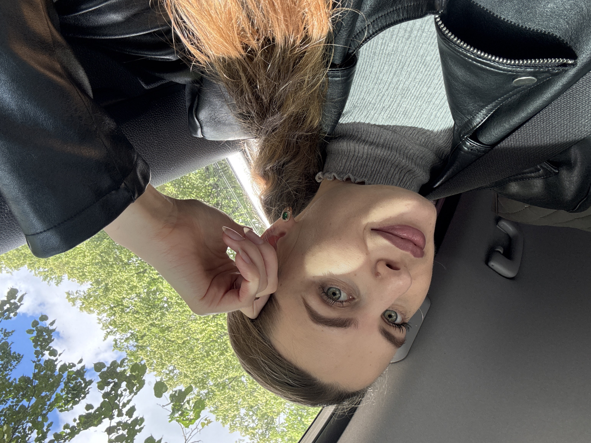 A young adult female is depicted in a close-up, taken from a slightly low angle inside a car. Her head is tilted, and she looks directly at the camera with a neutral expression. She has light-colored eyes, possibly green or hazel, and light to medium brown hair, styled back from her face. She is wearing a black leather jacket with visible zippers, over a grey, high-necked sweater with a ruffled edge. A small, green gemstone earring is visible in her right ear, and her nails appear manicured.
A black seatbelt diagonally crosses her chest, indicating she is a passenger in the vehicle. The interior of the car, including a dark grey ceiling or headliner, is visible around her. Through the car window, a bright outdoor scene unfolds, featuring vibrant green trees and foliage under a blue sky dotted with white clouds, suggesting a sunny daytime. The natural light casts shadows on her face. The image captures a moment of travel, likely a selfie taken by the woman, within the general context of Chaikovsky, Russia. No visible text is present in the image.