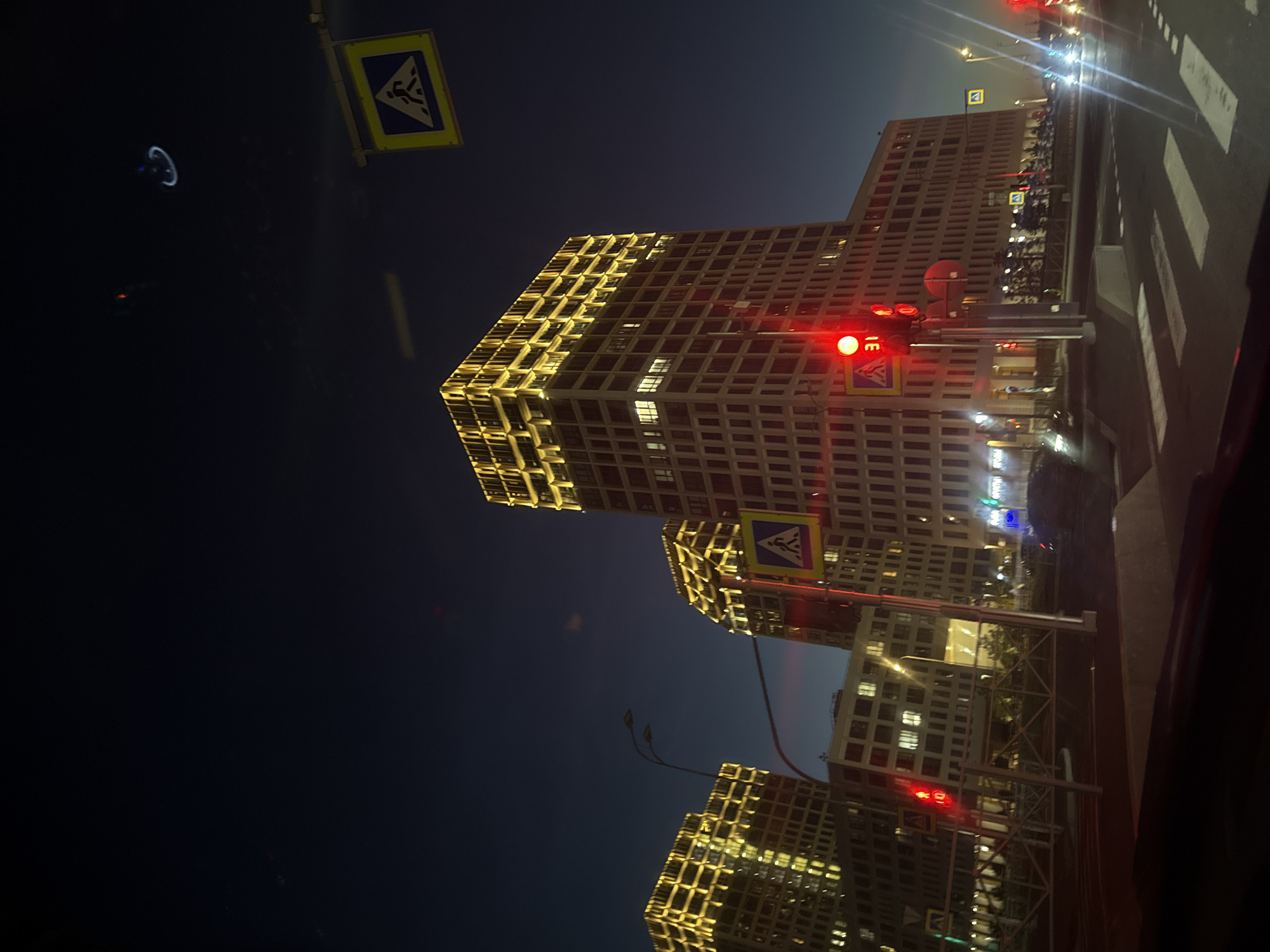 This image depicts an urban scene at dusk in Saint Petersburg, Russia. The foreground features a crosswalk with white painted lines on an asphalt road. Several cars are visible on the road, with their headlights on. Traffic lights are illuminated, with one red light prominent in the center of the frame. Two large, modern buildings, illuminated with warm yellow lights on their upper floors, dominate the background. These buildings appear to be residential or office complexes. Pedestrian crossing signs are also visible, mounted on poles near the road. The sky is a deep blue, indicating the transition from daylight to night. In the upper right quadrant of the sky, there is a faint, circular, blue and white light formation, possibly an aerial display or an artifact of the camera lens. The overall atmosphere is one of a bustling city at the end of the day.