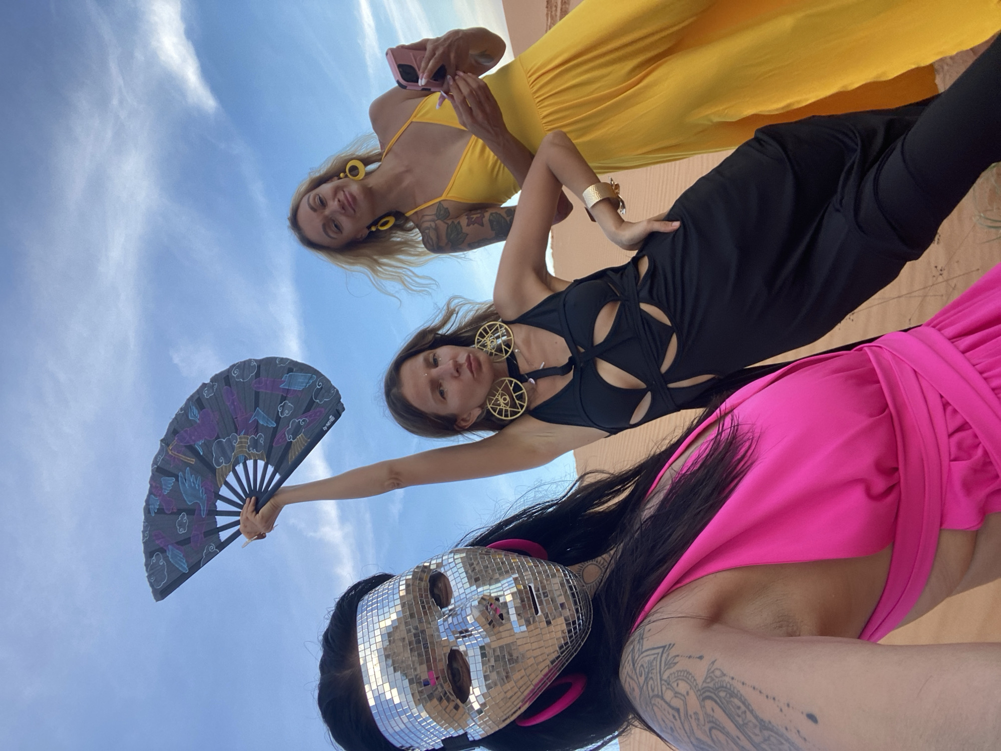 Three women pose for a selfie in a desert landscape. The woman in the foreground wears a mirrored mask and a bright pink dress. The woman in the middle wears a black dress with cutouts and holds a fan. The woman on the right wears a yellow dress and holds a phone. The background shows sand dunes and a cloudy blue sky.