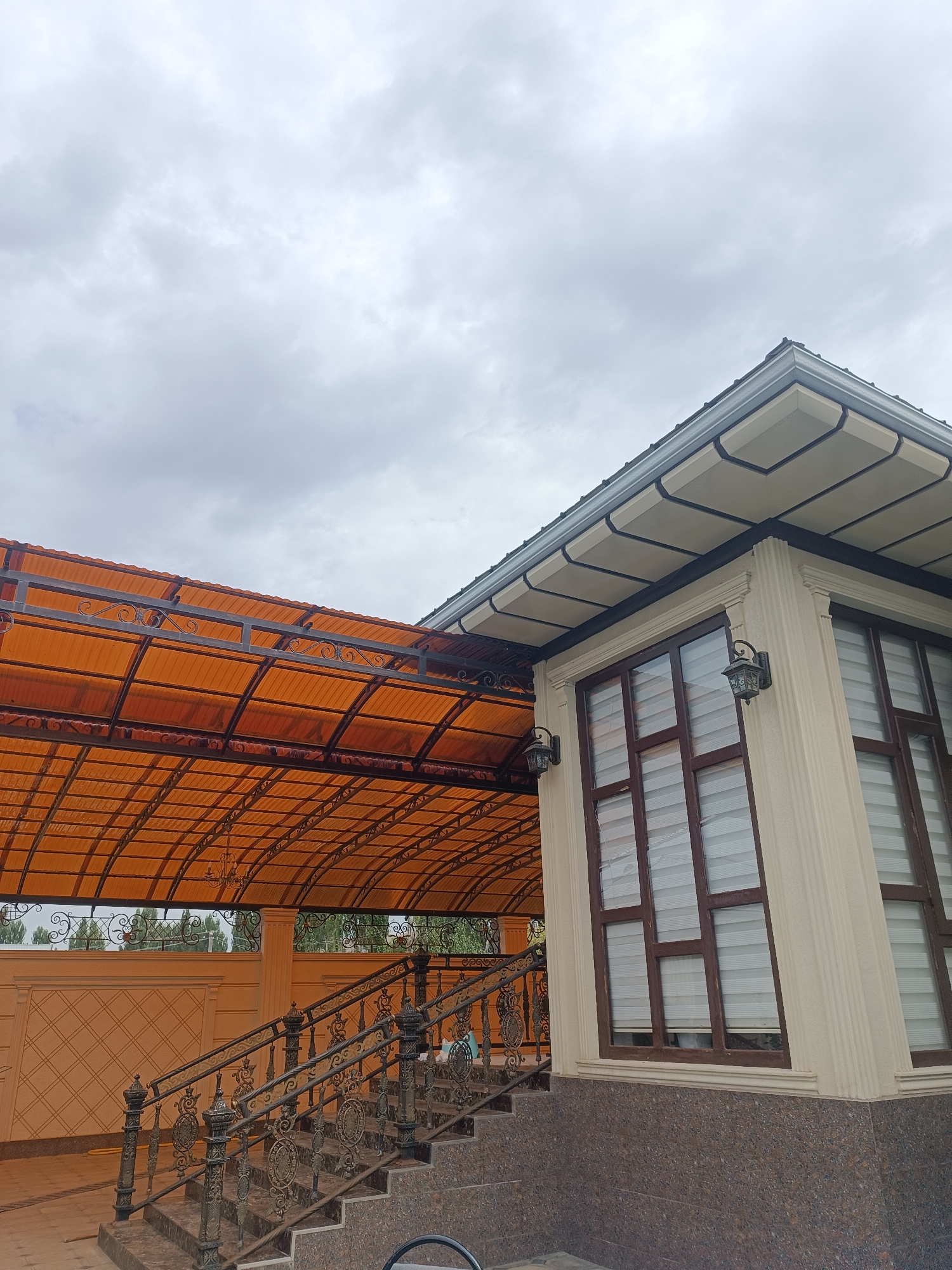 A low-angle outdoor shot captures a residential building with a distinct architectural style, likely in Myrza-Ake, Kyrgyzstan. The building features a light beige exterior with decorative fluted columns and a dark-rimmed roof with segmented decorative panels. Large windows with dark brown frames and white blinds are prominent. Two ornate, dark-colored wall sconces are attached to the building.
To the left, an orange, translucent awning extends from the building, supported by a dark metal frame with decorative scrollwork. The awning is curved and covers a set of stairs with ornate dark metal railings. The stairs are made of speckled brown material. Behind the stairs and awning, a light beige wall with a diamond pattern is visible.
The sky above is overcast with light gray clouds, suggesting a potentially cool or rainy day. The overall scene depicts a well-maintained property with a focus on decorative elements, particularly in the metalwork of the awning and railings. There are no people visible in the frame.