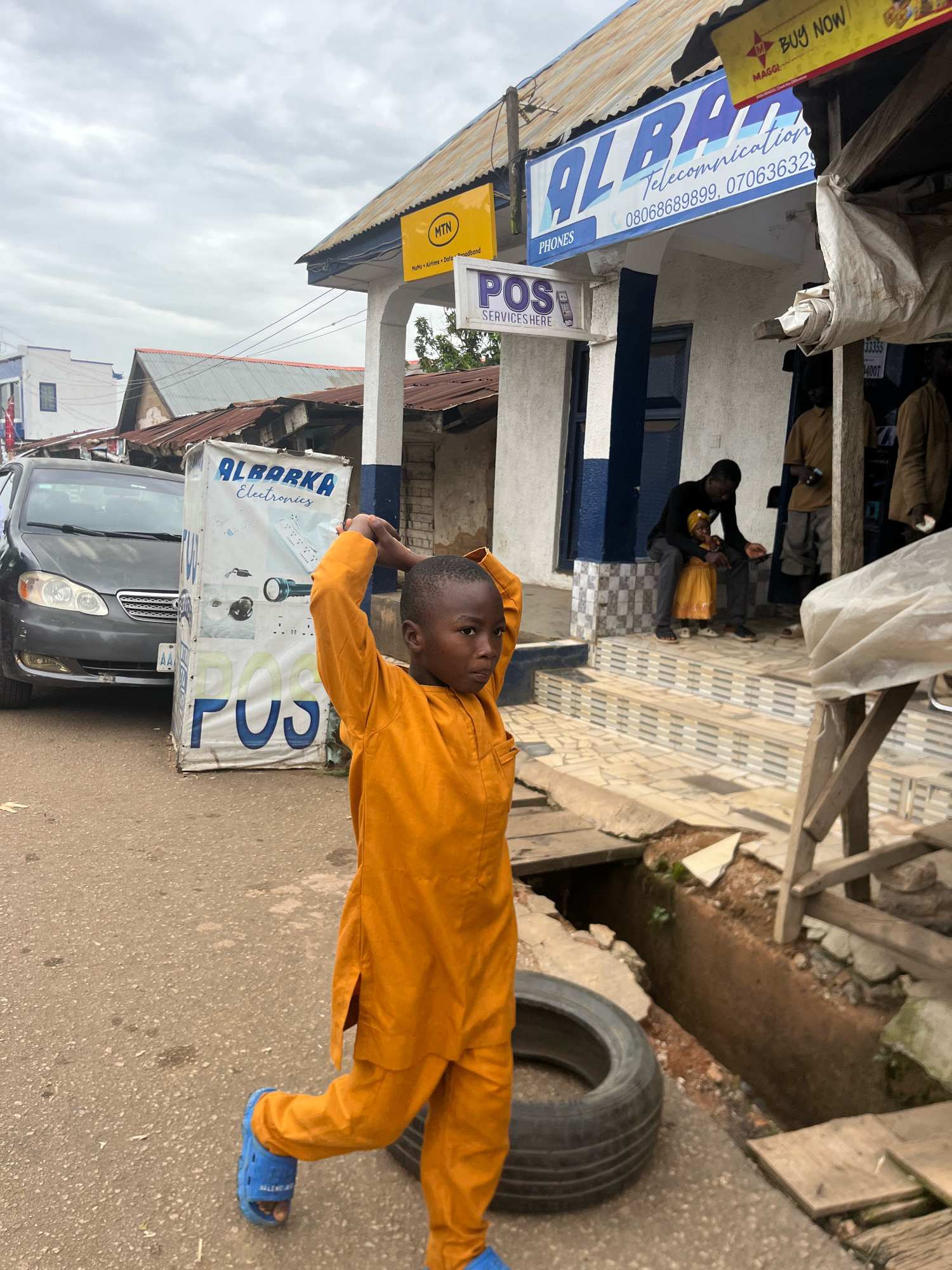 In Jos, Nigeria, a young boy wearing an orange outfit and blue sandals stands on a paved street in front of a building with signs for "ALBARKA Telecommunication" and "POS SERVICES HERE". To the left, a dark grey sedan is parked. A large, white, rectangular advertising stand with "ALBARKA Electronics" and "POS" written on it is positioned next to the boy. The building's exterior is painted white with blue trim. Stairs lead up to the building entrance, where two individuals are visible. One person is seated with a child on their lap, while another stands nearby. The scene appears to be an outdoor commercial area under an overcast sky, with some debris and a tire visible on the ground.