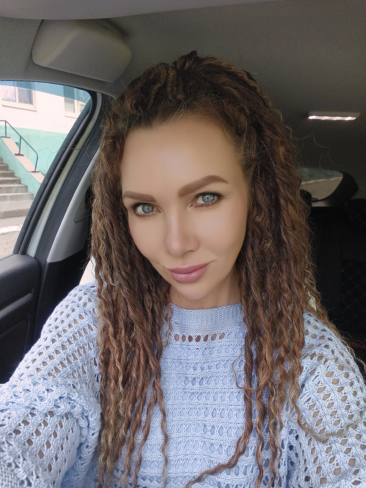A woman with curly, brown hair is seated in a car. She is wearing a light blue, knitted sweater with a patterned openwork design. The car is parked with a view of a building with a staircase outside the window. The staircase has white steps and a green railing. The location appears to be within a city, but the specific city and region are not discernible from the image.