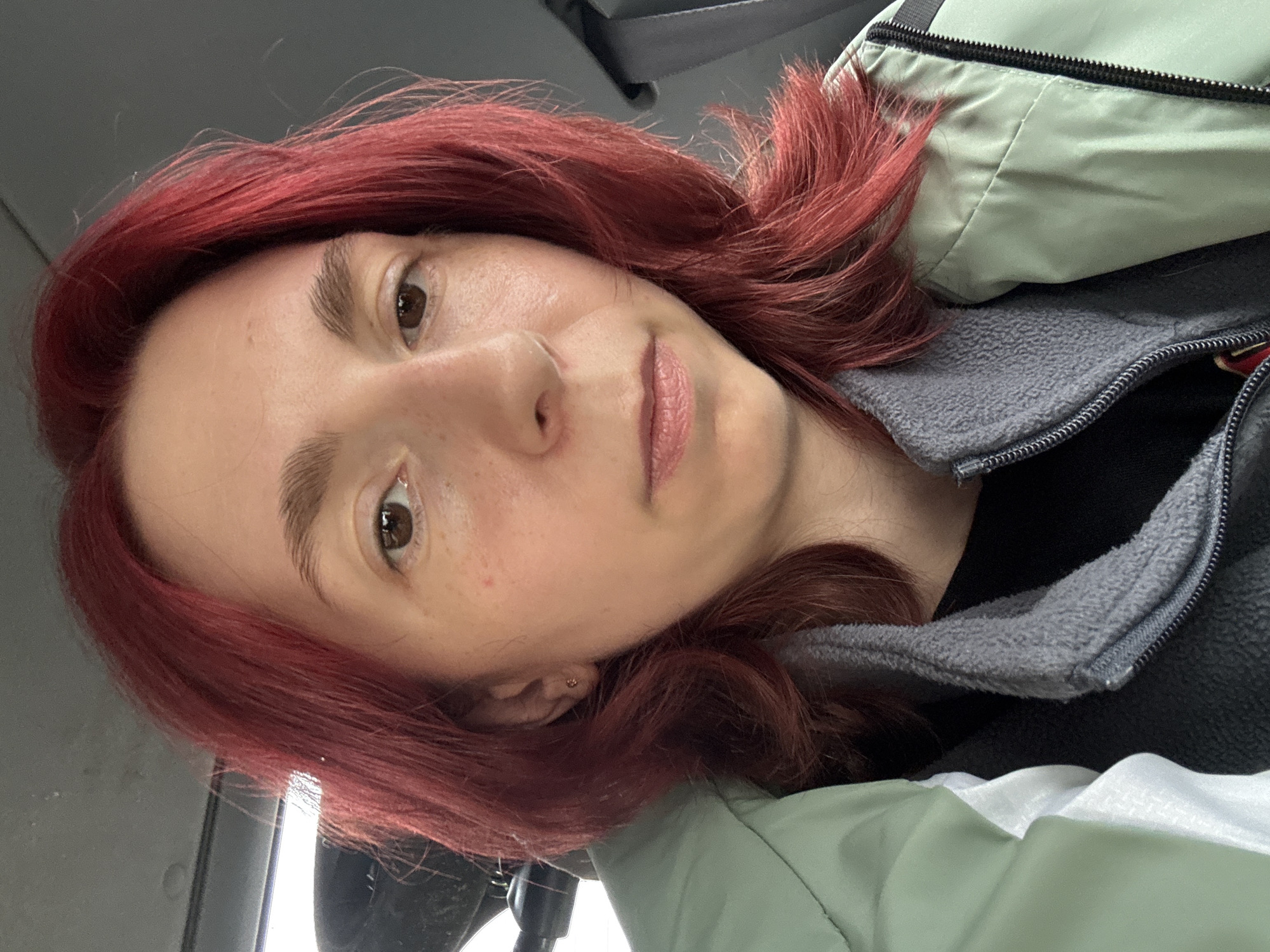 A medium-shot, eye-level, indoor selfie shows a young Caucasian woman with bright red hair and fair skin. She is looking directly at the camera, with a neutral expression. The woman is wearing a light green jacket over a black shirt and a gray fleece collar. Her hair is shoulder-length and wavy, with some strands falling forward to frame her face. She has noticeable freckles across her cheeks and nose. The background is blurred and appears to be the interior of a vehicle, possibly a car, with the headliner and a seatbelt visible. The lighting suggests an overcast day or a dimly lit indoor environment.