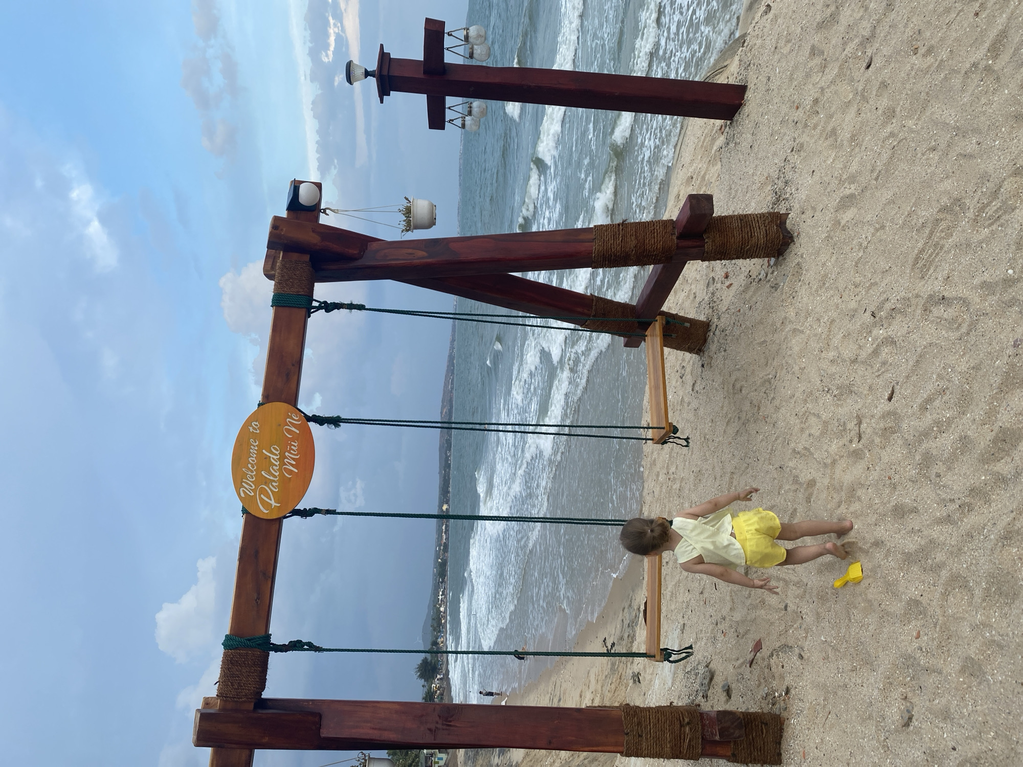 A child in a yellow outfit is on a swing at Palado Mui Ne Beach in Phu Thuy Ward, Vietnam. The swing set is made of dark wood and rope. The sign reads "Welcome to Palado Mui Ne." The beach has sand and the ocean is in the background. There are also wooden posts with lights. The sky is cloudy.