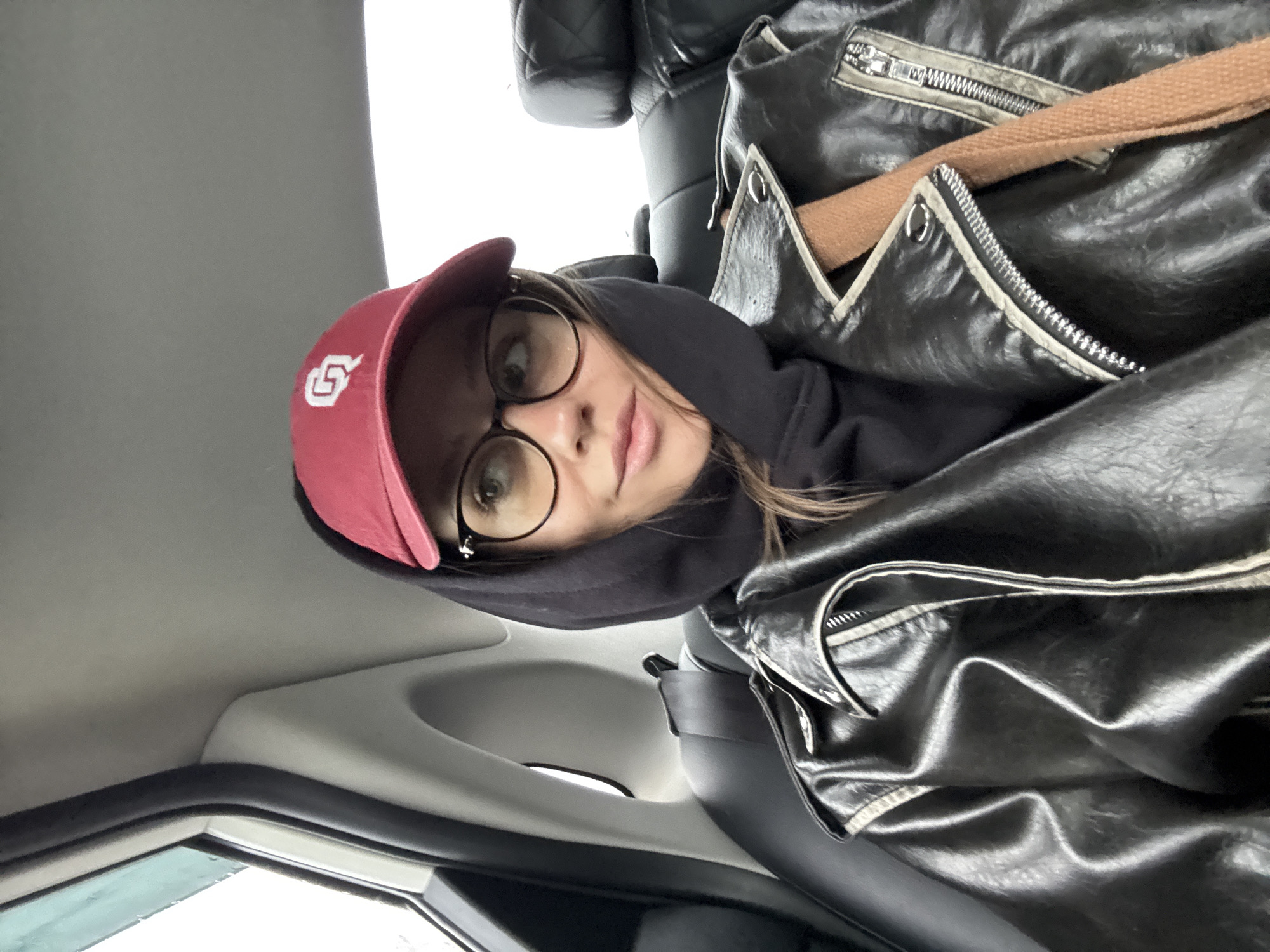 A woman with glasses and a red baseball cap is in a car. She is wearing a black hoodie and a black leather jacket. The interior of the car is gray and black. The location is unknown, but it is likely not Krasnoyarsk, Russia due to the absence of indicators within the image.