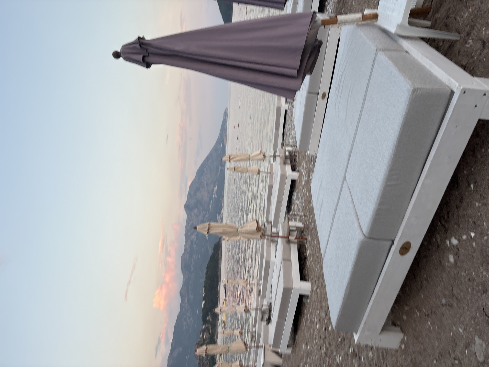 The image captures a serene beach scene in Budva, Montenegro, at dusk. The focus is on a row of white beach chairs with gray cushions, each accompanied by closed beige umbrellas. These chairs are positioned along a pebble beach, with the calm, shimmering sea extending into the distance. In the background, mountains rise, partly obscured by the fading light and some clouds. The sky is a mix of pale blue and pink, suggesting the time is around sunset or early evening. There are no visible people or specific actions taking place in the shot.