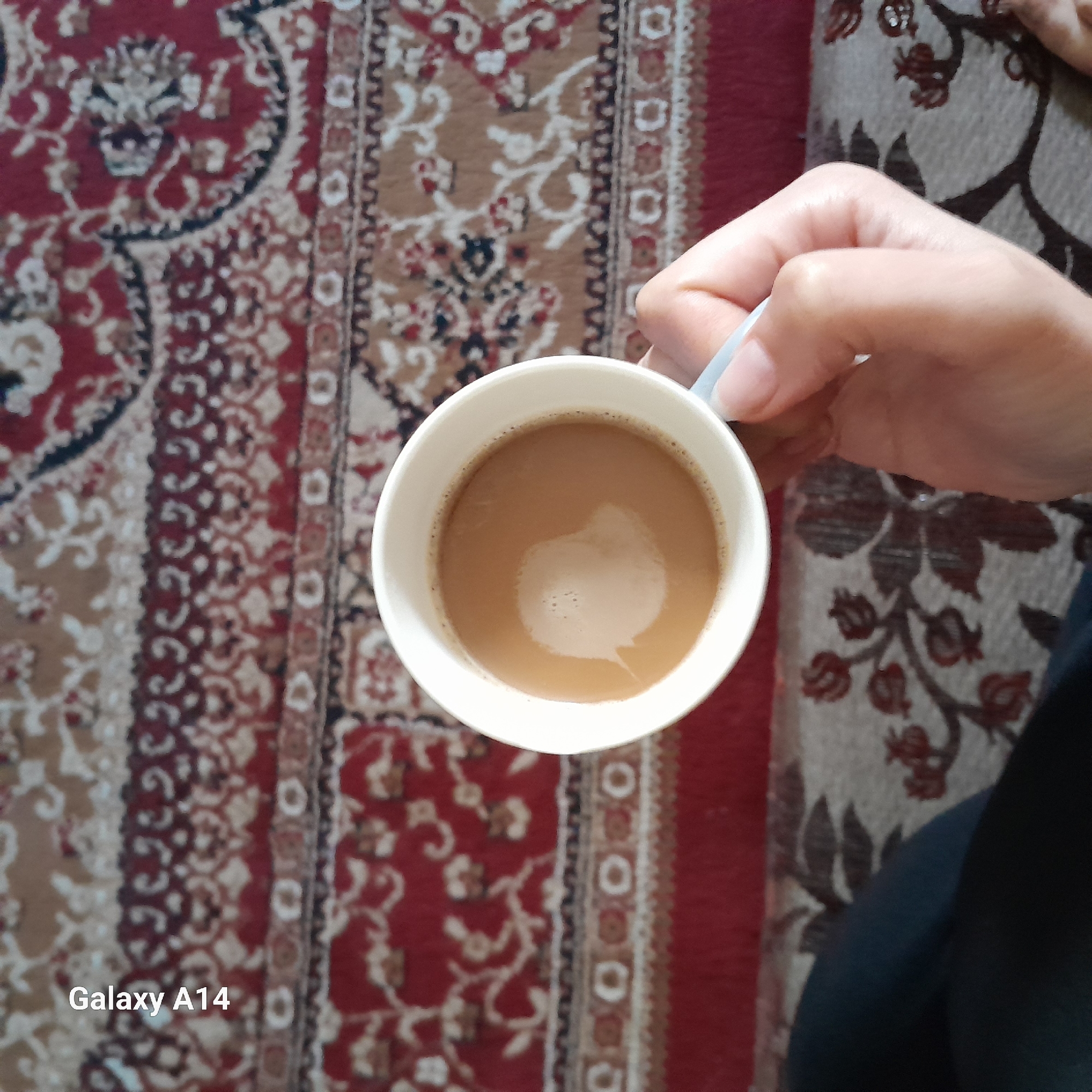 A photograph depicts a white cup of coffee held by a hand. The cup is positioned over a patterned carpet. The camera brand Galaxy A14 can be seen on the bottom left. The location is Unknown, Unknown.