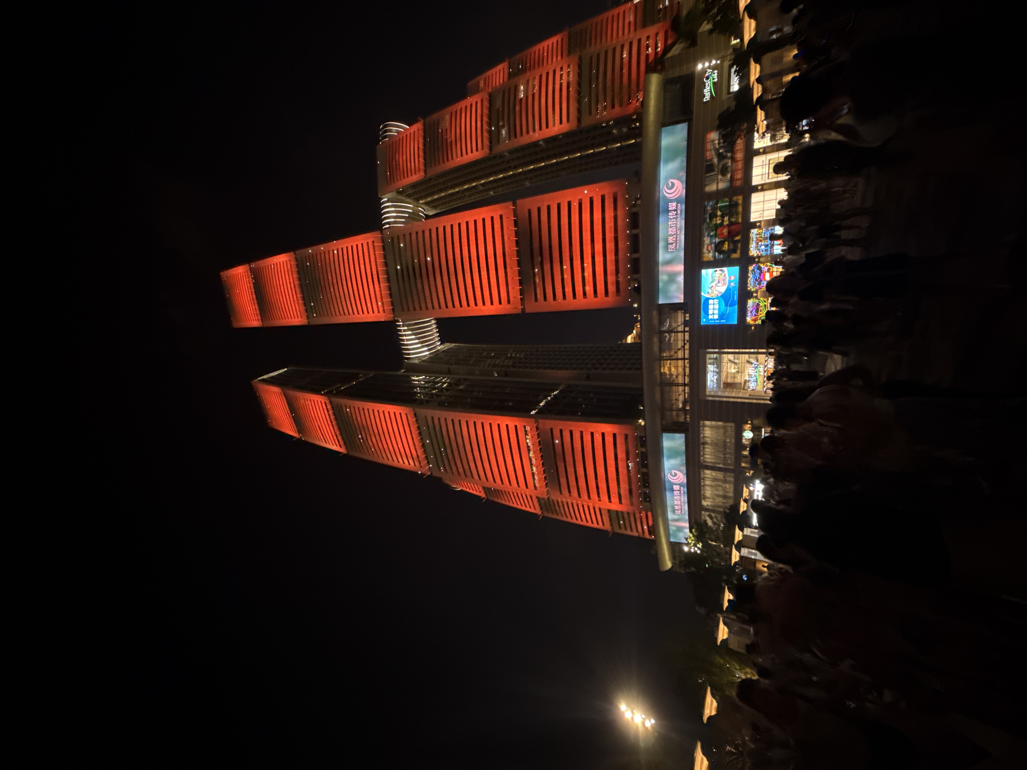 A low-angle, night-time shot captures the Raffles City complex in Chongqing, Yuzhong District, China. The iconic twin skyscrapers, illuminated in vibrant red, dominate the frame against a dark sky. The buildings are characterized by horizontal lines of red light interspersed with darker bands, suggesting architectural features. A horizontal connecting structure links the two main towers at a significant height.
Below the towers, a multi-story building forms the base of the complex, featuring large digital billboards. One billboard displays Chinese characters and a logo with a swirling design. Another billboard shows colorful animated content. The lower section of this building has large glass windows and appears to be a commercial space, with the "Raffles City" logo visible on the right.
In the foreground, a crowd of people is silhouetted against the illuminated buildings and billboards. They are gathered in a public space, likely a plaza or street, in front of the complex. The figures are indistinct due to the low light and motion, with some appearing to be holding up phones or cameras. The overall scene suggests a lively urban environment with a focus on modern architecture and public gathering. To the left of the frame, a bright, multi-bulb light fixture is visible, adding to the urban illumination.