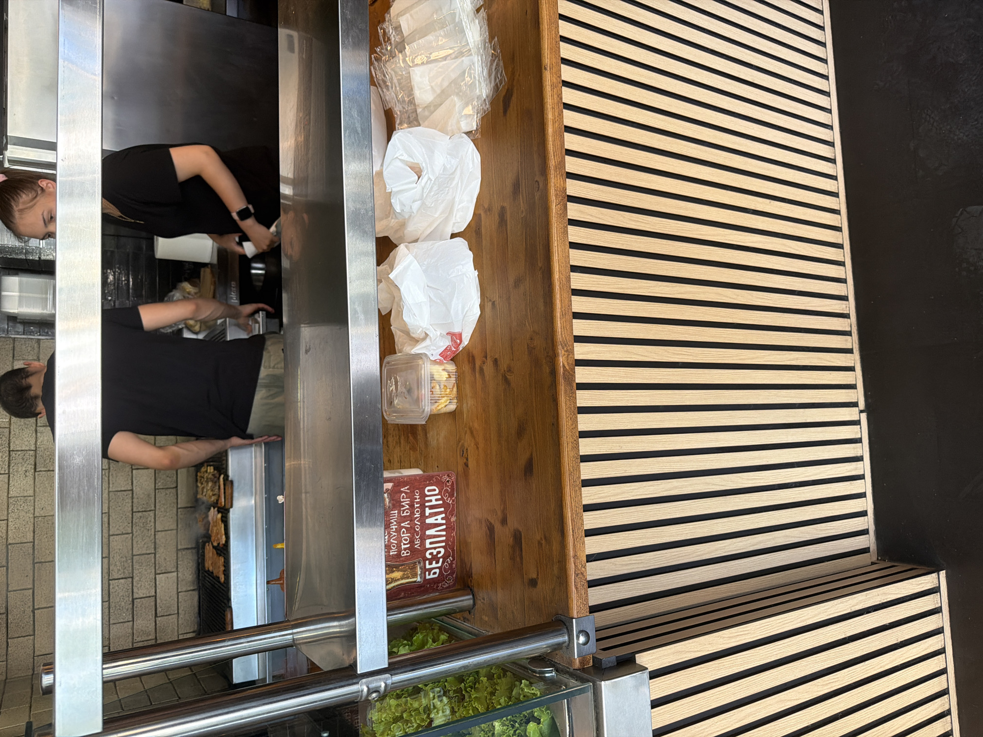 The image depicts a food service counter with two employees, a man and a woman, working behind it. The man, wearing a black t-shirt and light-colored pants, is standing to the left, facing away from the camera, and appears to be attending to a grill. The woman, wearing a black t-shirt and a necklace, is to the right, also facing away from the camera, and is holding a white object. The counter displays various items, including a sign in Cyrillic script, plastic containers, and white paper bags. To the left of the counter, a glass display case contains fresh green lettuce. The background features a tiled wall and stainless steel shelving. The overall scene suggests a bustling food establishment. The question about the city of Basarbovo, Bulgaria, cannot be answered from the image itself, as no identifying location markers are visible.