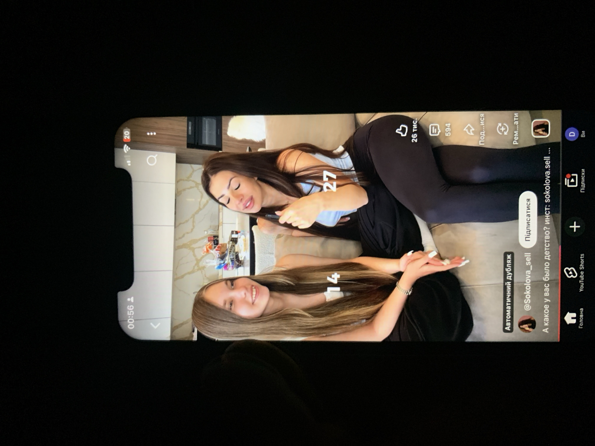 In the image, two young women are seated on a couch, likely indoors given the kitchen counter visible in the background. The woman on the left, with long, light brown hair, smiles gently. The woman on the right, with dark brown hair, is holding a phone or similar device and appears to be speaking. Both are wearing casual clothing. A digital overlay on the screen shows "00:56" and other interface elements, suggesting this is a recording or playback on a mobile device. Below the women, text and icons indicate social media or video platform content, with user information "@Sokolova_sell" visible. The scene is presented in portrait orientation on a screen, which is then photographed, resulting in a dark border around the content. There is no visible indication of the location being Zhmerynka, Ukraine, nor any elements that specifically place it in that city.