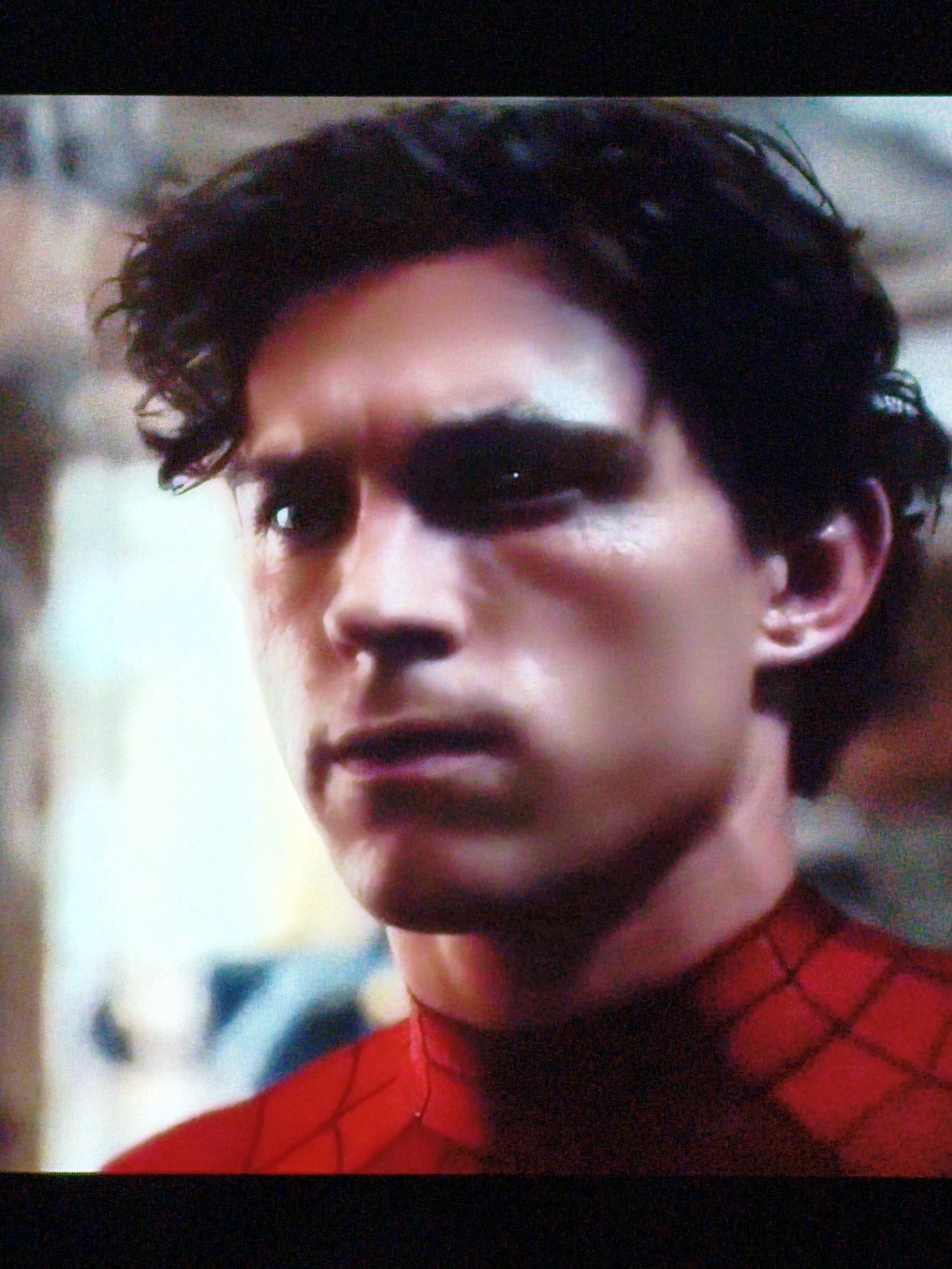 The image shows a close-up, slightly grainy, headshot of a young man with dark, curly hair. He is wearing a red suit with a black web pattern, characteristic of Spider-Man's costume. The man's face is partially illuminated, revealing his features and the texture of his skin, which shows some signs of wear or injury on his left cheek. His expression is neutral or somber. The background is out of focus and appears to be an indoor or possibly an urban environment with muted colors and textures, suggesting a film or television still. There is no discernible connection to Vladivostok, Russia, as the image provides no geographical cues.