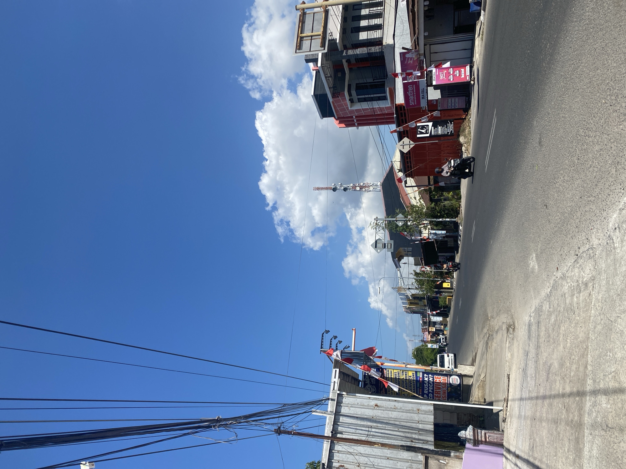 The image depicts a street scene in Bulukumba, Indonesia. The sky is bright blue with scattered white clouds. Several power lines are visible in the upper left portion of the image. The street is paved and appears to be a main thoroughfare with buildings lining both sides. On the left side, there is a corrugated metal building with signage and what appears to be a purple-painted pillar. Small Indonesian flags are displayed on poles along the sidewalk. On the right side of the street, various commercial buildings are present, including one with a prominent red and white facade and another with a "smartfren" advertisement. A cell tower is visible in the background. Traffic includes a white van and a motorcycle with a rider. The overall environment suggests a commercial or residential street under a sunny sky.