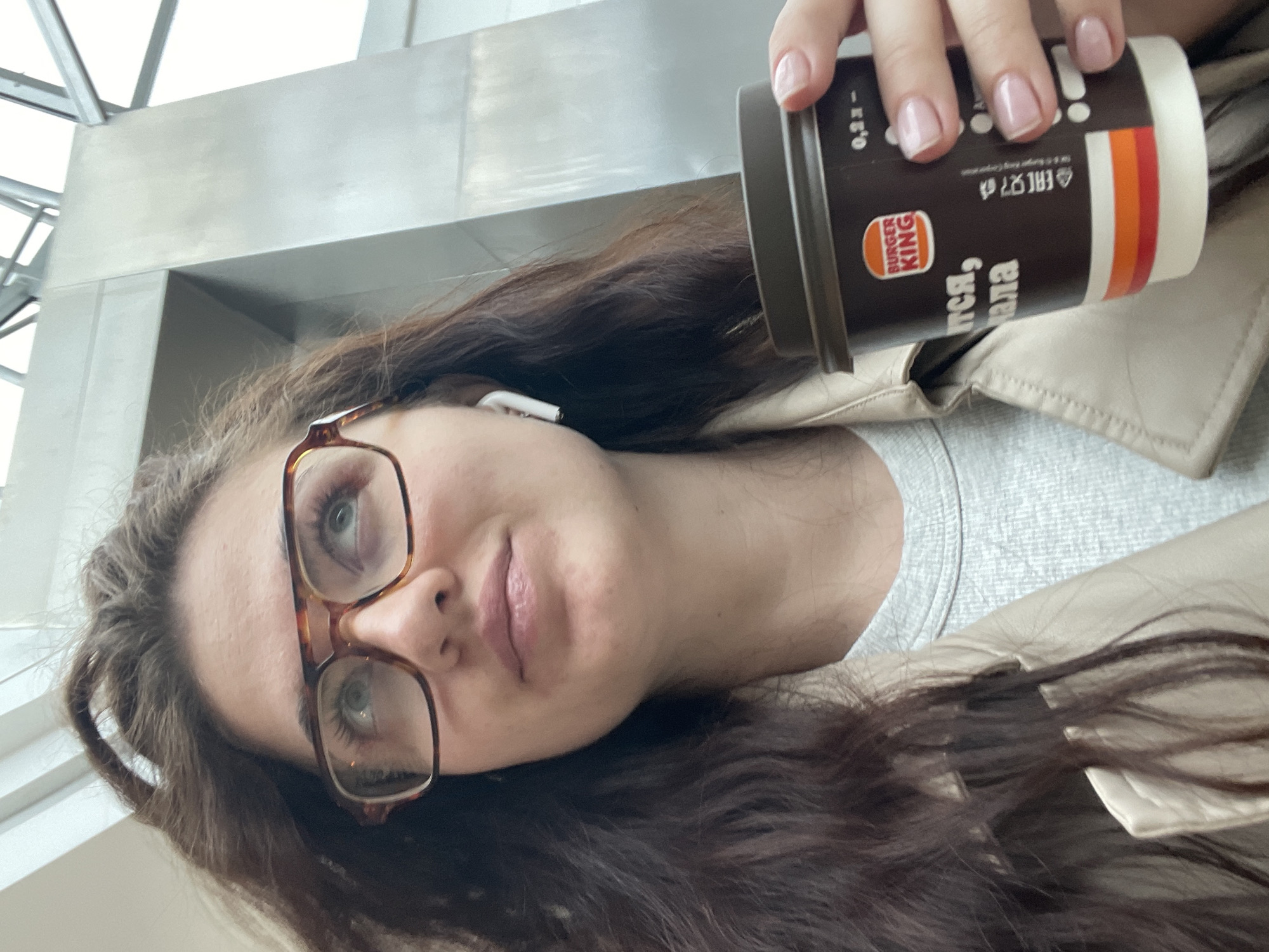 The image shows a woman wearing glasses and an earbud. She is holding a Burger King coffee cup with Russian text. The background suggests an airport setting, potentially in Moscow, Russia, based on the Cyrillic script on the coffee cup. The woman appears to be taking a selfie.