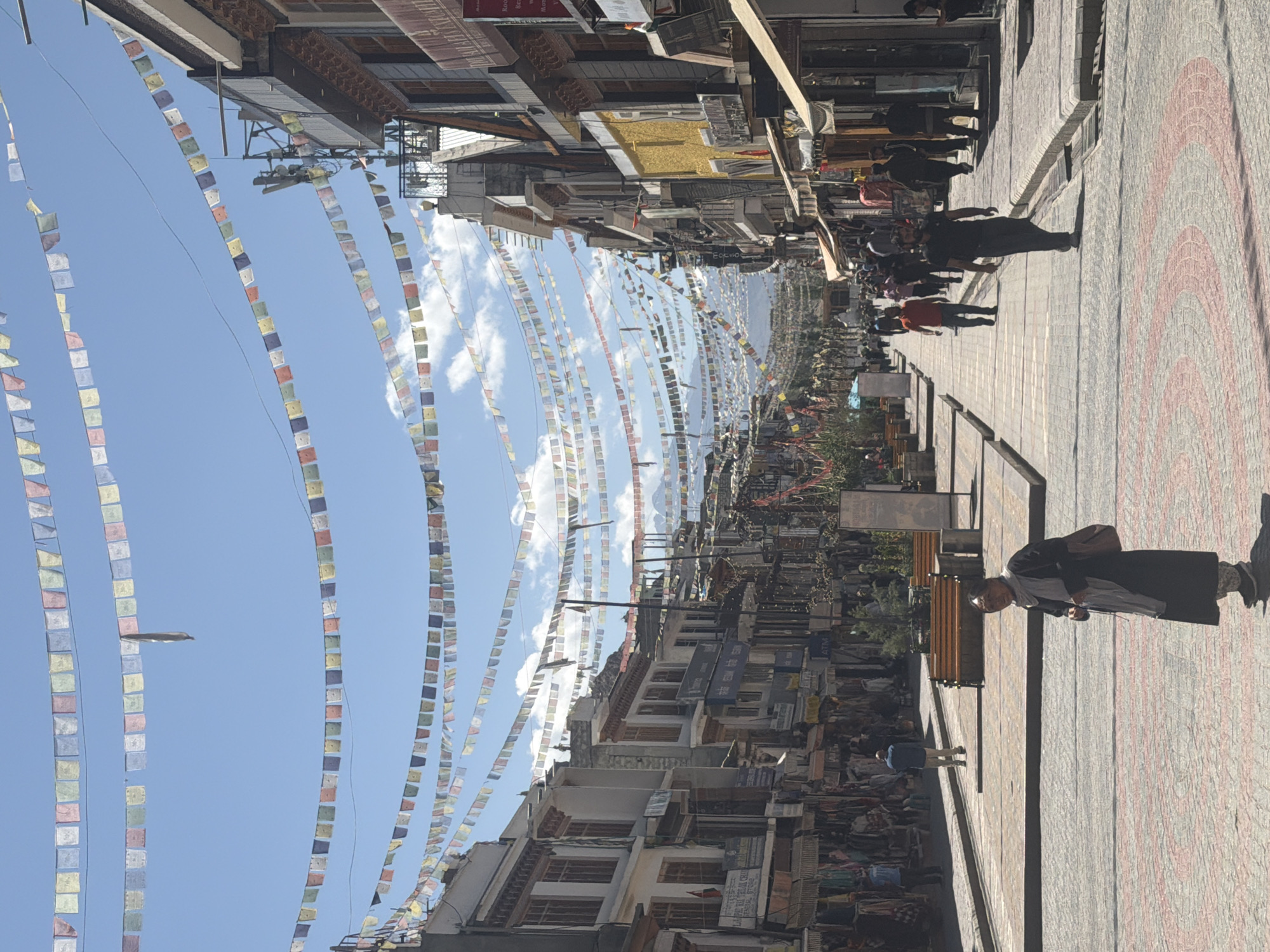 The image depicts a bustling street scene in the city of Leh, India, likely on the main market road. The street is lined with traditional Ladakhi architecture, featuring multi-story buildings with wooden accents and balconies. Colorful prayer flags are strung across the street at various heights, creating a vibrant canopy against the clear blue sky with scattered clouds.
A variety of people are present, walking along the street and browsing shops. In the foreground, an elderly woman in traditional attire, a dark robe and a grey scarf, is walking towards the viewer. She carries a bag over her shoulder. Further down the street, men and women in casual clothing are seen. Some are walking purposefully, while others appear to be shopping or socializing. The street is paved with interlocking bricks forming a decorative pattern. Shops and stalls line both sides of the street, displaying goods like textiles and handicrafts. Benches are placed along the sidewalk for rest. In the background, mountains are visible, indicating the mountainous terrain of Ladakh. The overall atmosphere is one of a lively and active urban environment.