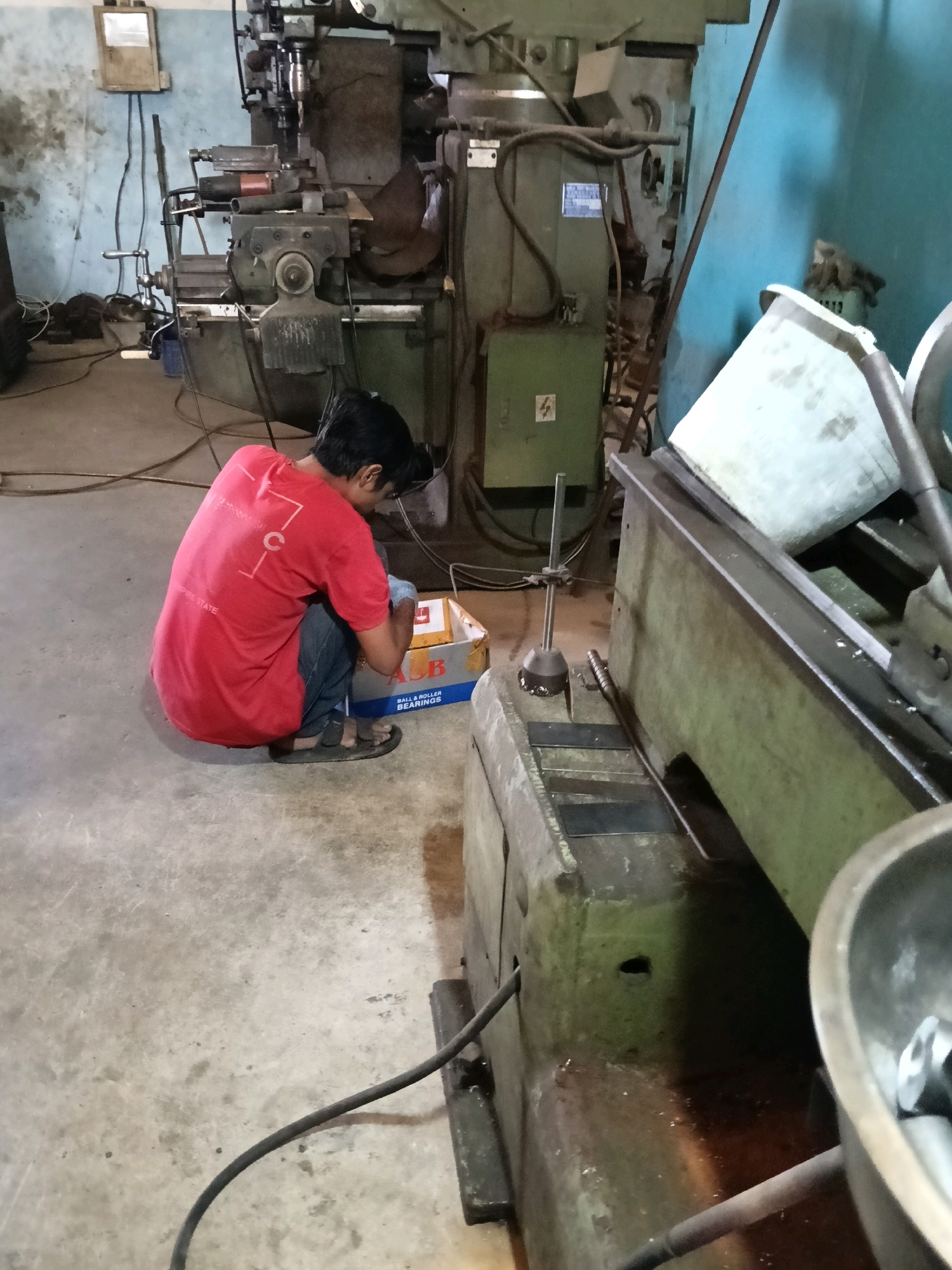 In a dimly lit workshop in Belimbing, Indonesia, a young person wearing a red t-shirt crouches on a concrete floor, facing a cardboard box. The box, partially open, reveals what appears to be industrial equipment. The workshop is filled with large, complex machinery, including a milling machine and other metalworking equipment, suggesting a manufacturing or repair facility. The environment appears functional but somewhat cluttered, with wires and tools scattered around. The person is focused on the box, possibly inspecting or organizing its contents. The overall scene conveys an atmosphere of industrial work and productivity.