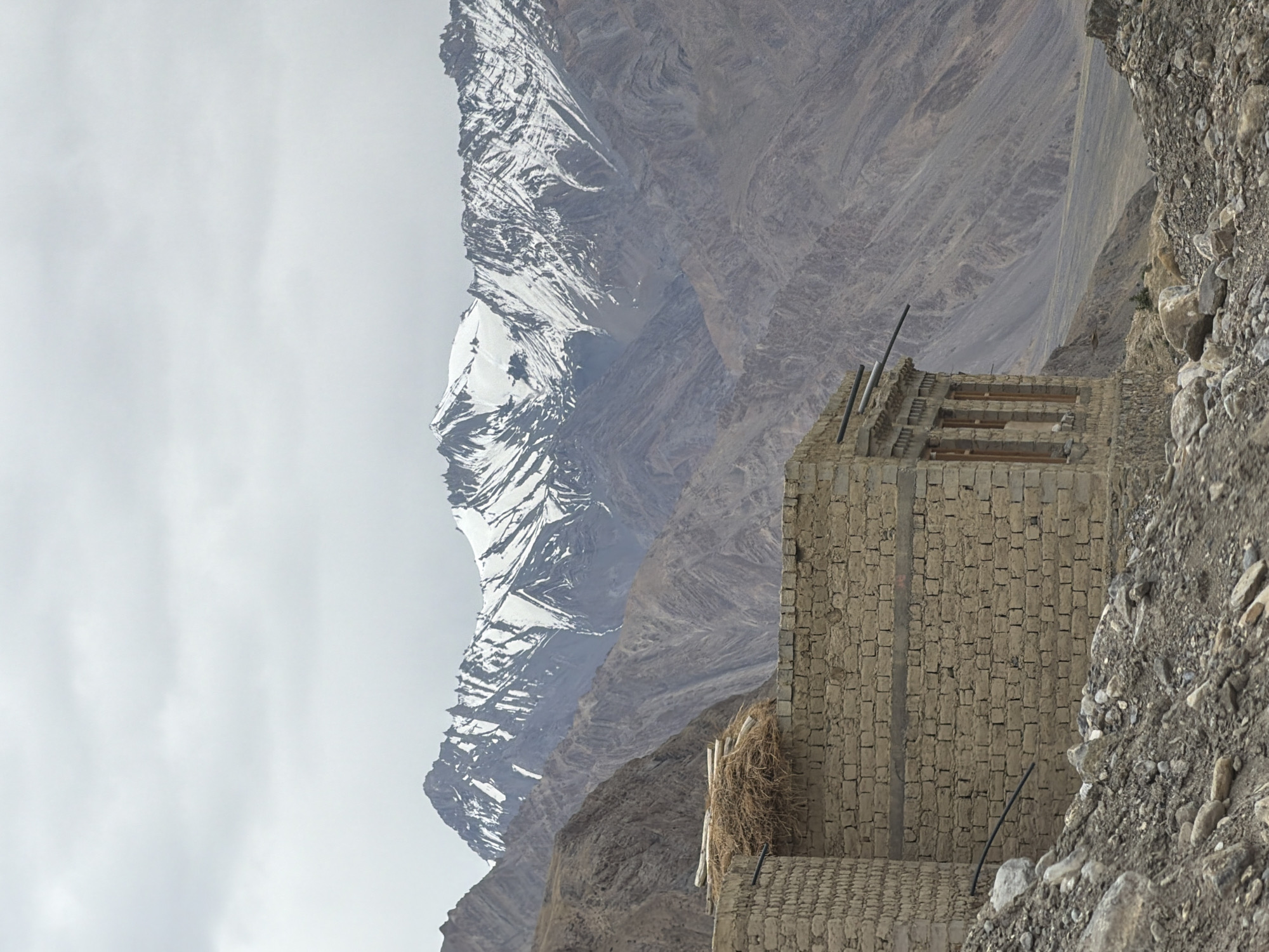 An overcast, cloudy sky hangs over a rugged landscape in Zangla, India. In the foreground, a partially constructed building made of stone bricks stands incomplete. A pile of straw and wooden beams is situated on top of one section of the wall. The building's windows are visible, some with wooden frames. A black pipe extends from the building into the surrounding terrain. The ground leading up to the structure is littered with rocks and debris. In the mid-ground, steep, barren mountains rise, their slopes textured with striations and erosion. In the background, a majestic mountain range is visible, its peaks capped with snow, creating a stark contrast with the arid lower slopes. The overall scene depicts a remote, mountainous environment with signs of human habitation in an undeveloped state.