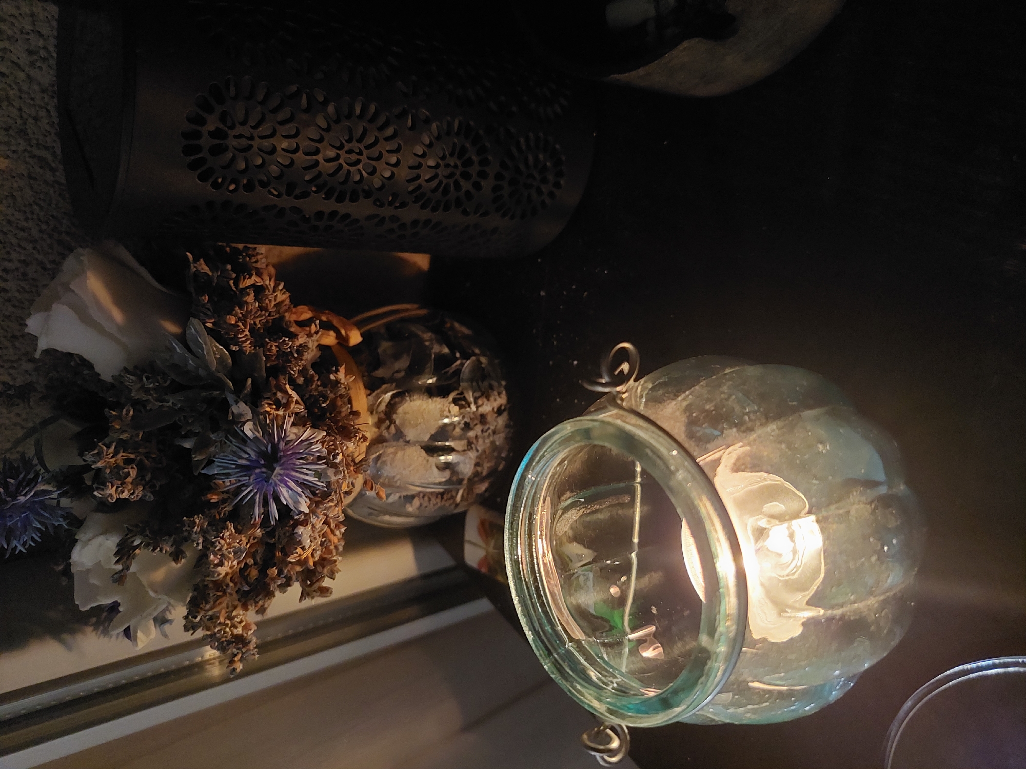 The image shows an indoor still life arrangement. A dark-colored, cylindrical object with decorative perforations sits at the top left of the frame, partially obscuring a floral arrangement of dried and artificial flowers, including white and purple blooms, positioned below it. To the right, two glass vessels are visible. One is a spherical, patterned glass container, and the other, in the foreground, is a clear, textured glass lantern with a metal rim, containing a light source. The scene is dimly lit, with the light source illuminating the immediate area around the lantern. The setting appears to be a flat surface. There is no indication of Plovdiv, Bulgaria, or any other specific location.