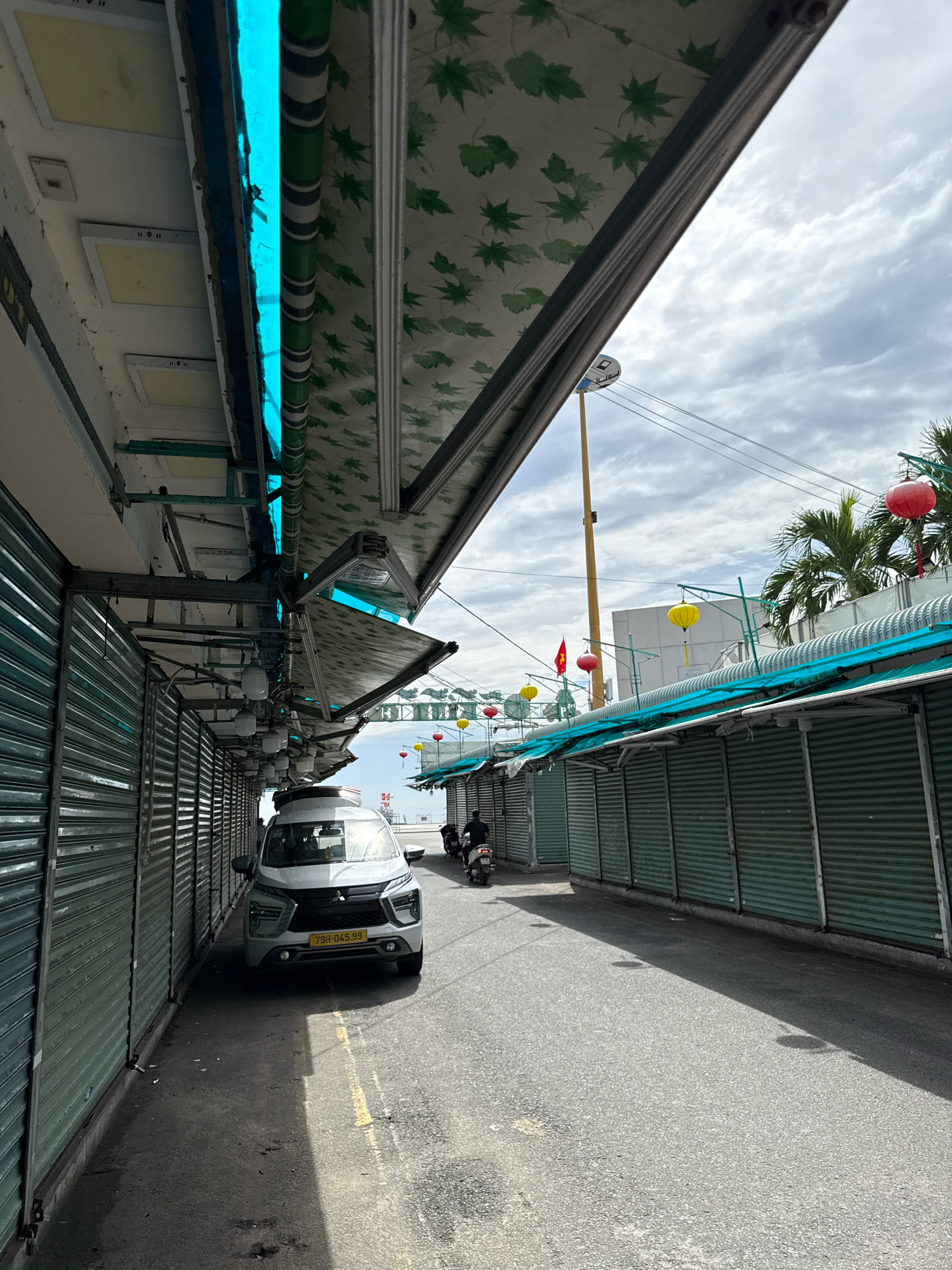 The image shows a street scene in Nha Trang, Vietnam, likely in a market area. The perspective is low, looking down a narrow street flanked by closed market stalls with corrugated metal shutters. A light-colored SUV, identified by its license plate "79H-045.99", is parked on the left side of the street. The stalls are covered by awnings, one of which is brightly lit with blue neon lights. Green awnings with a leaf pattern are visible on some stalls. Red and yellow lanterns are strung across the street in the background, suggesting a festive or commercial setting. Palm trees are visible behind some of the stalls. The sky is partly cloudy. Two motorcycles are present; one is parked near the SUV, and another is being ridden away from the camera down the street. The overall impression is of a quiet period, possibly early morning or a time when the market is closed.
