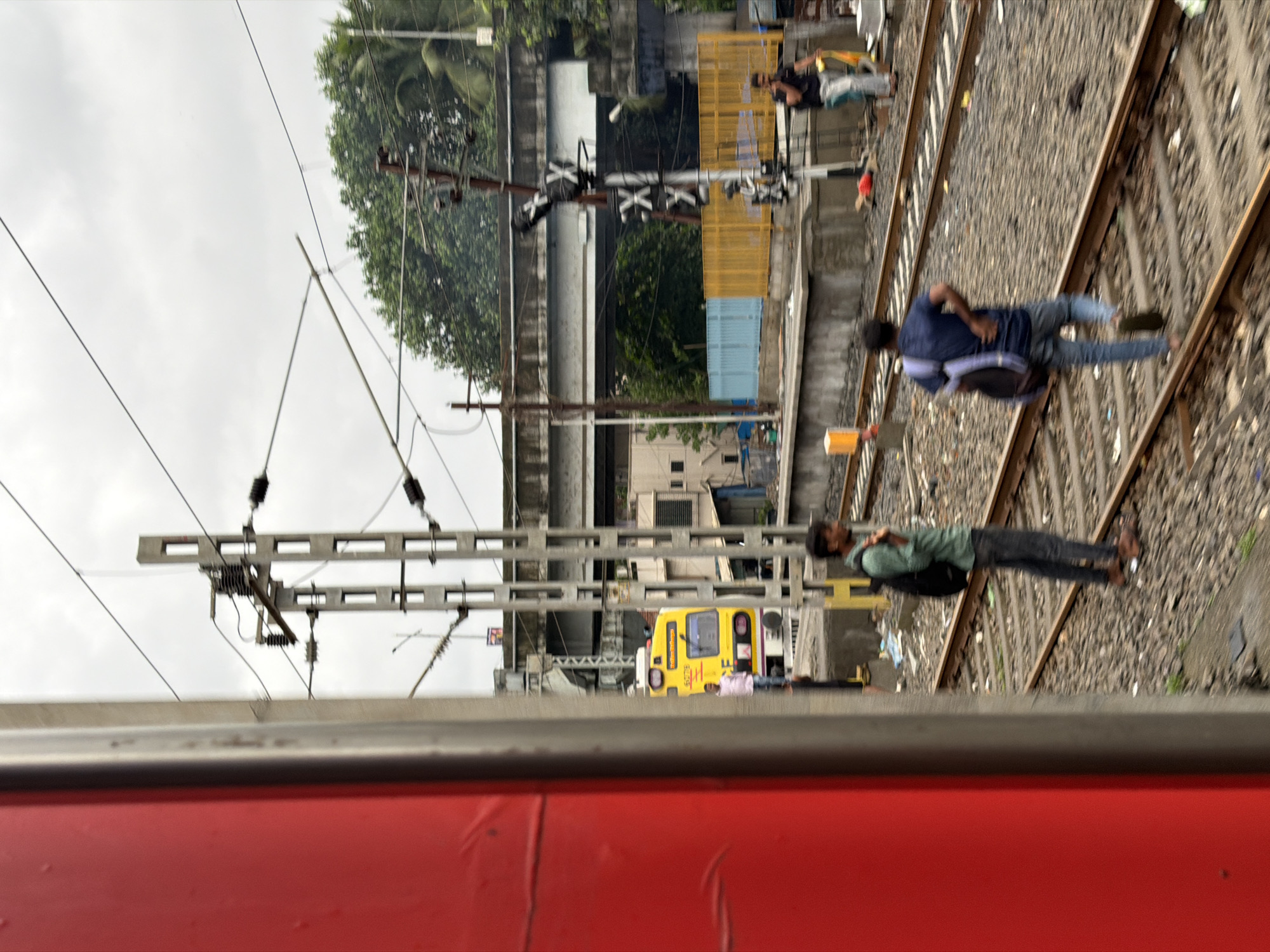 The image captures a scene at the Vasai Road railway station in Vasai-Virar, India. On the left, a portion of a red train carriage is visible, blurred due to motion. In the background, a yellow local train is parked at the platform. Several individuals are present on or near the tracks and platform. One man in a green shirt and dark pants is standing by the tracks, carrying a backpack. Another man in a blue shirt and jeans is walking away from the camera on the tracks. Further in the background, a person in a black shirt and light pants is standing on the platform. Overhead power lines and signal posts are prominent features of the railway environment. The scene appears to be under an overcast sky.