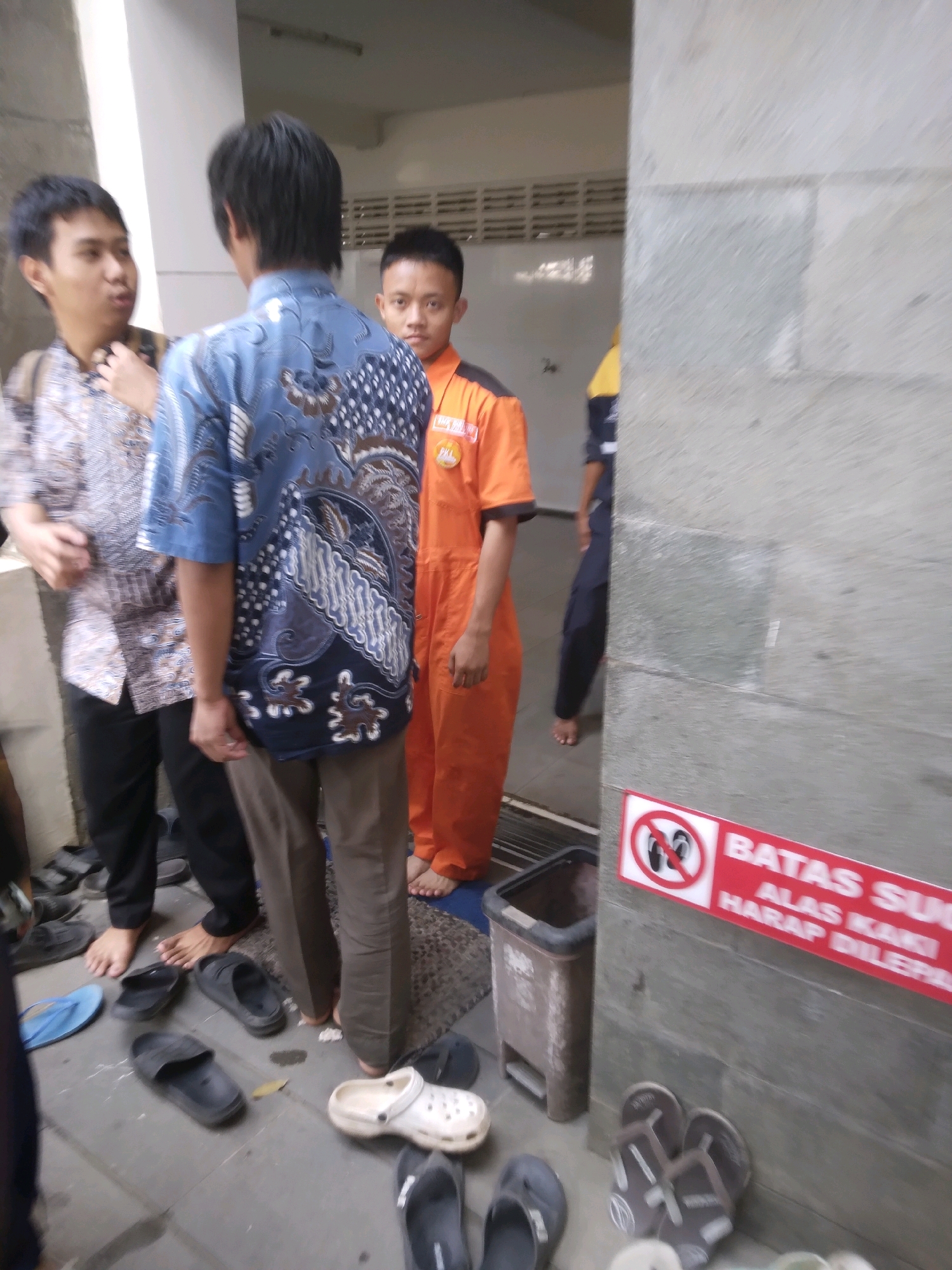 The image depicts a group of four young men standing in what appears to be an indoor communal space, possibly a hallway or entrance. The men are casually dressed, with some wearing patterned shirts and others in work uniforms. Two men are facing away from the camera, engaged in conversation. One is wearing a blue shirt with a batik pattern, and the other is wearing an orange jumpsuit. A third man, wearing a patterned shirt, is facing the viewer, and a fourth man, in an orange uniform, is partially visible on the right side of the frame. The floor is tiled and scattered with various footwear, including sandals and flip-flops. A red and white sign with text and a prohibition symbol is visible on a grey textured wall on the right. A trash bin is also present in the scene. The environment suggests a functional, everyday setting in Sokaraja, Indonesia.