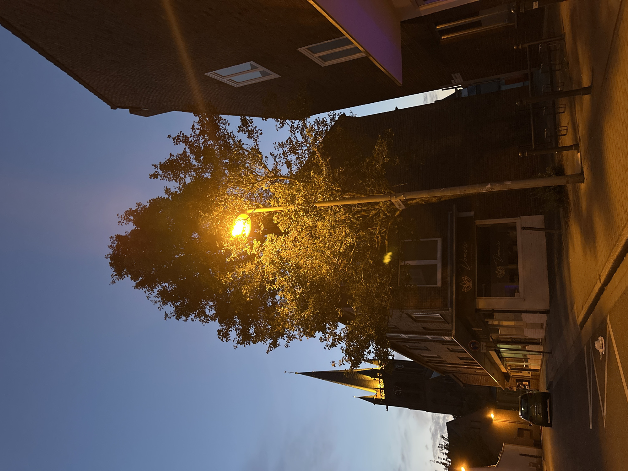 The image depicts a street scene in Krefeld, Germany, during dusk or early evening. A large tree, illuminated by a streetlamp, is prominent in the foreground, its leaves casting a warm glow. To the left, a church tower with a spire and clock is visible, also lit up. Several brick buildings line the street, with the one on the right featuring modern architecture and a purple-lit awning. A dark-colored car is parked on the left side of the street, and a trash can is situated on the sidewalk. The sky is a clear, deep blue, transitioning to a lighter hue near the horizon. A street sign reads "Krugerstr."